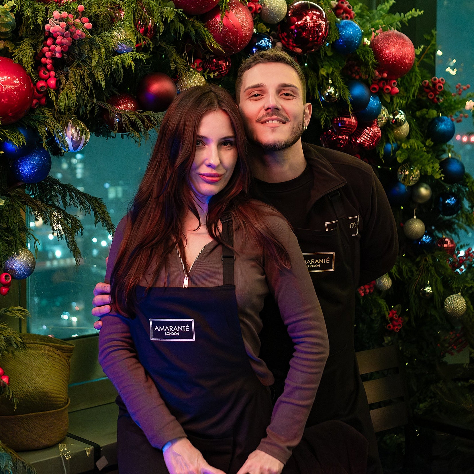 Two people wearing Amarante London aprons smiling in front of a festive Christmas wreath display during a luxury wreath-making workshop in London.