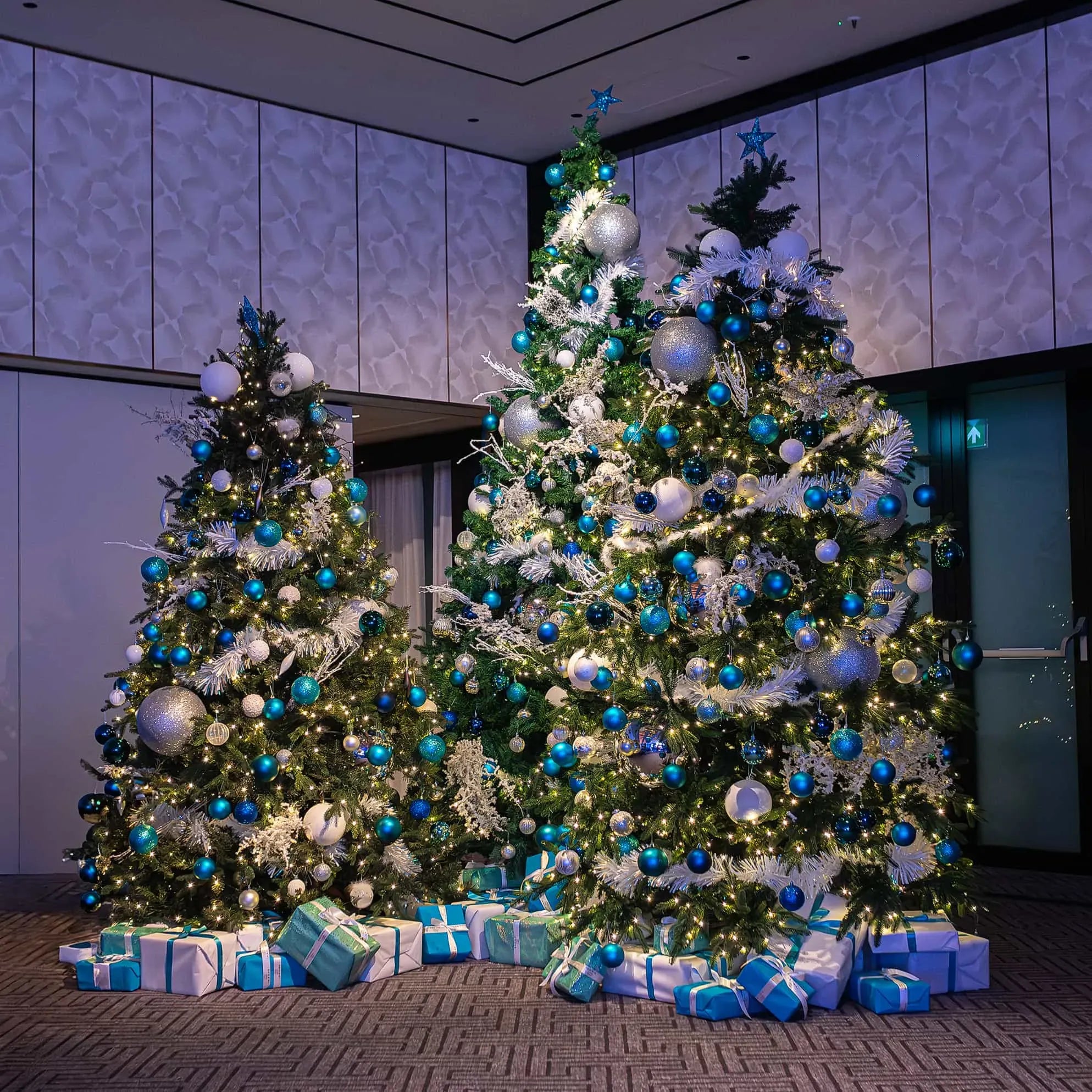 Two large Christmas trees decorated with white and blue ornaments, white lights, and topped with silver stars, surrounded by wrapped gifts in blue and silver paper, in a modern room with textured white walls and vertical light fixtures.