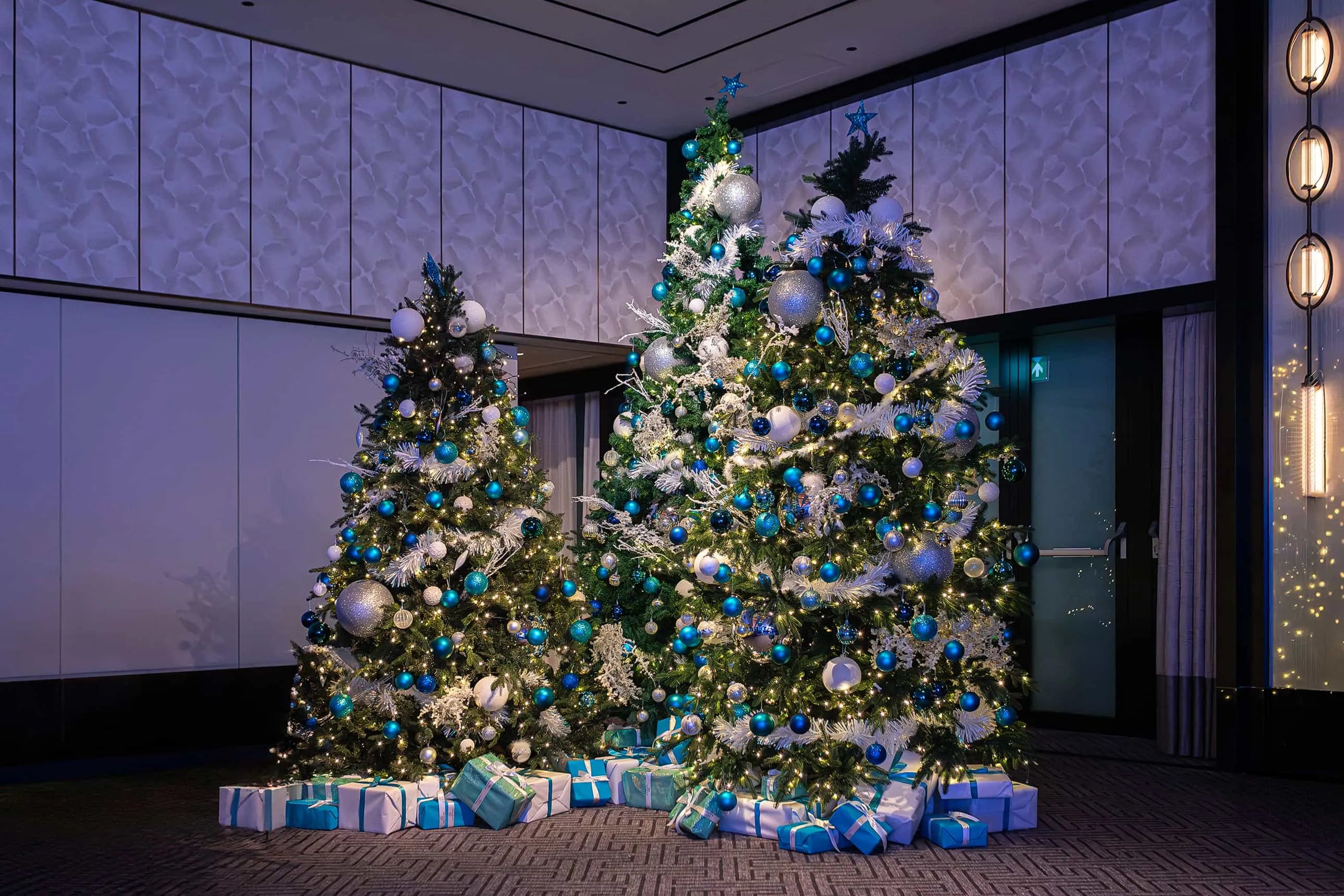 Two large Christmas trees decorated with white and blue ornaments, white lights, and topped with silver stars, surrounded by wrapped gifts in blue and silver paper, in a modern room with textured white walls and vertical light fixtures.