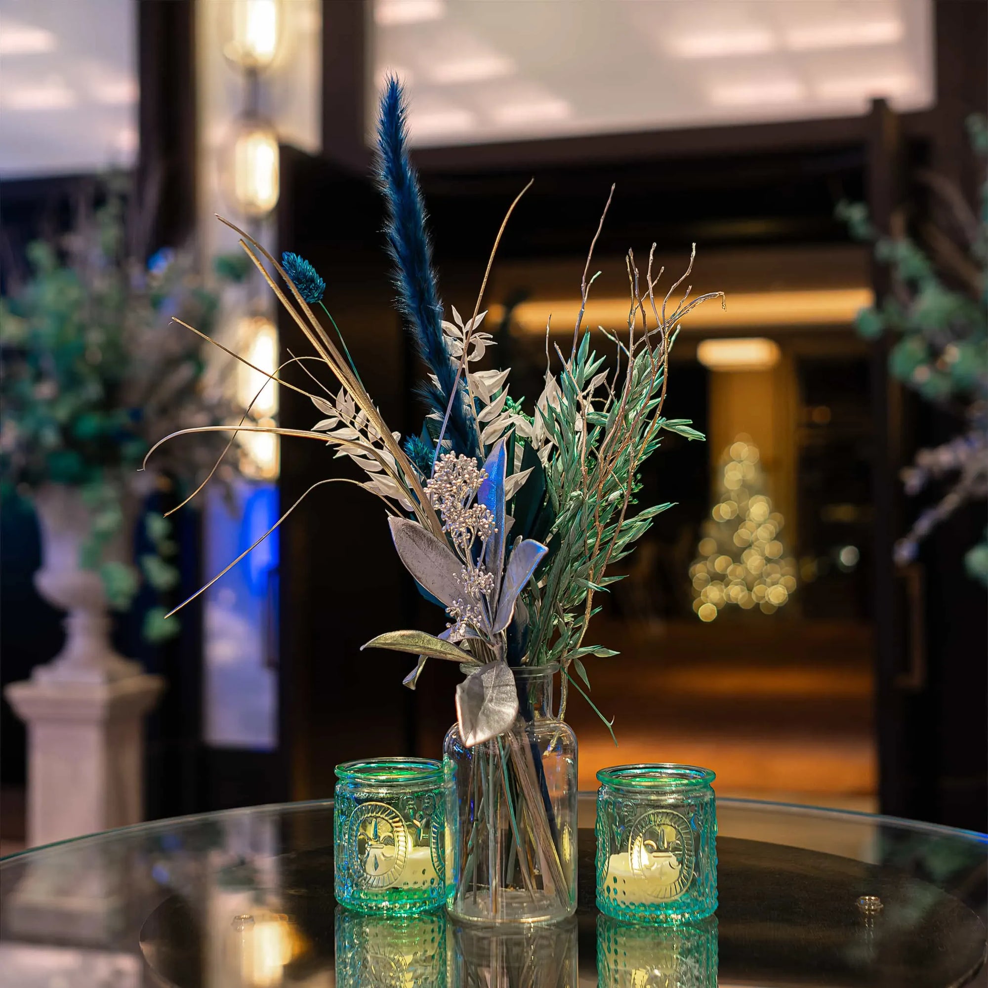 A tabletop with Christmas Flowers, a delicate composition of silver and blue foliage in a vase, including a soft feather accent, flanked by two glowing teal candle holders, set against the warm ambience designed by Amaranté London for the InMode event at Nobu Hotel.