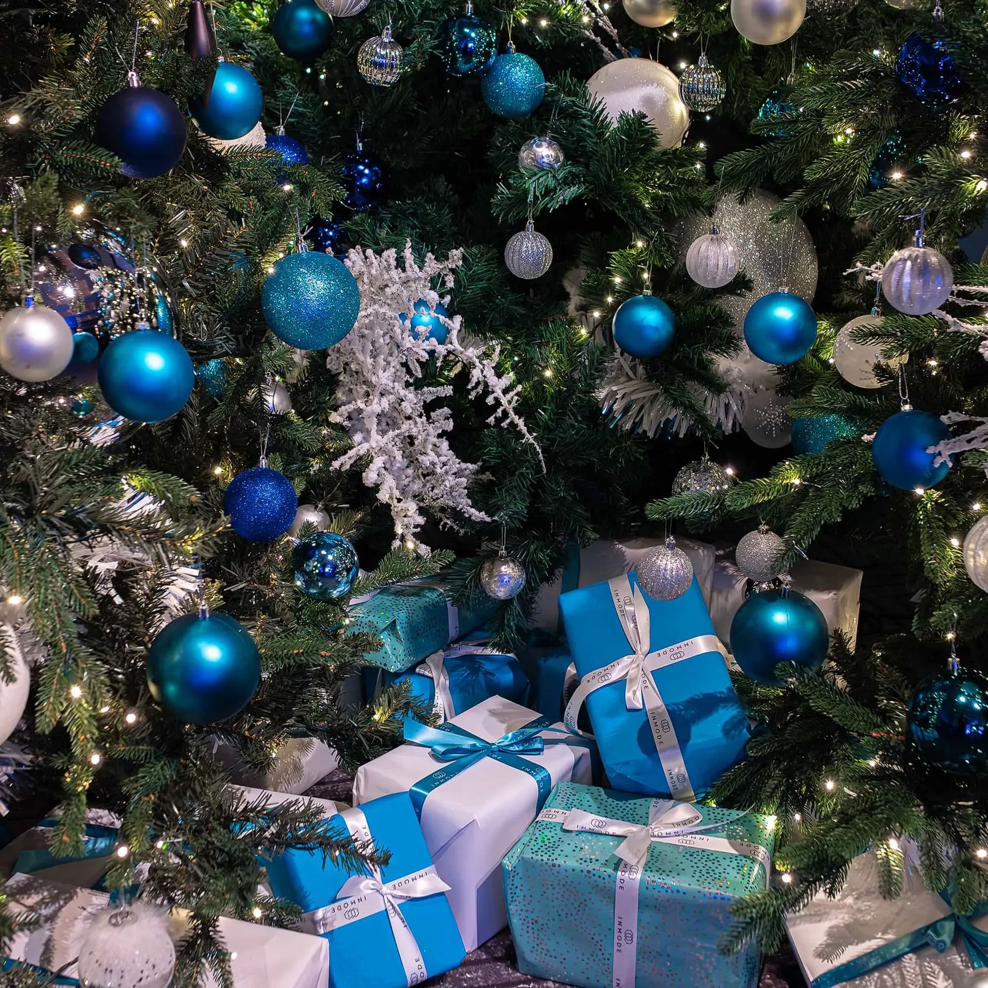 Details of a A Christmas tree richly adorned with blue and silver baubles, sparkling lights, and white decorative snowflakes, surrounded by a collection of presents wrapped in blue and white paper with ribbons.