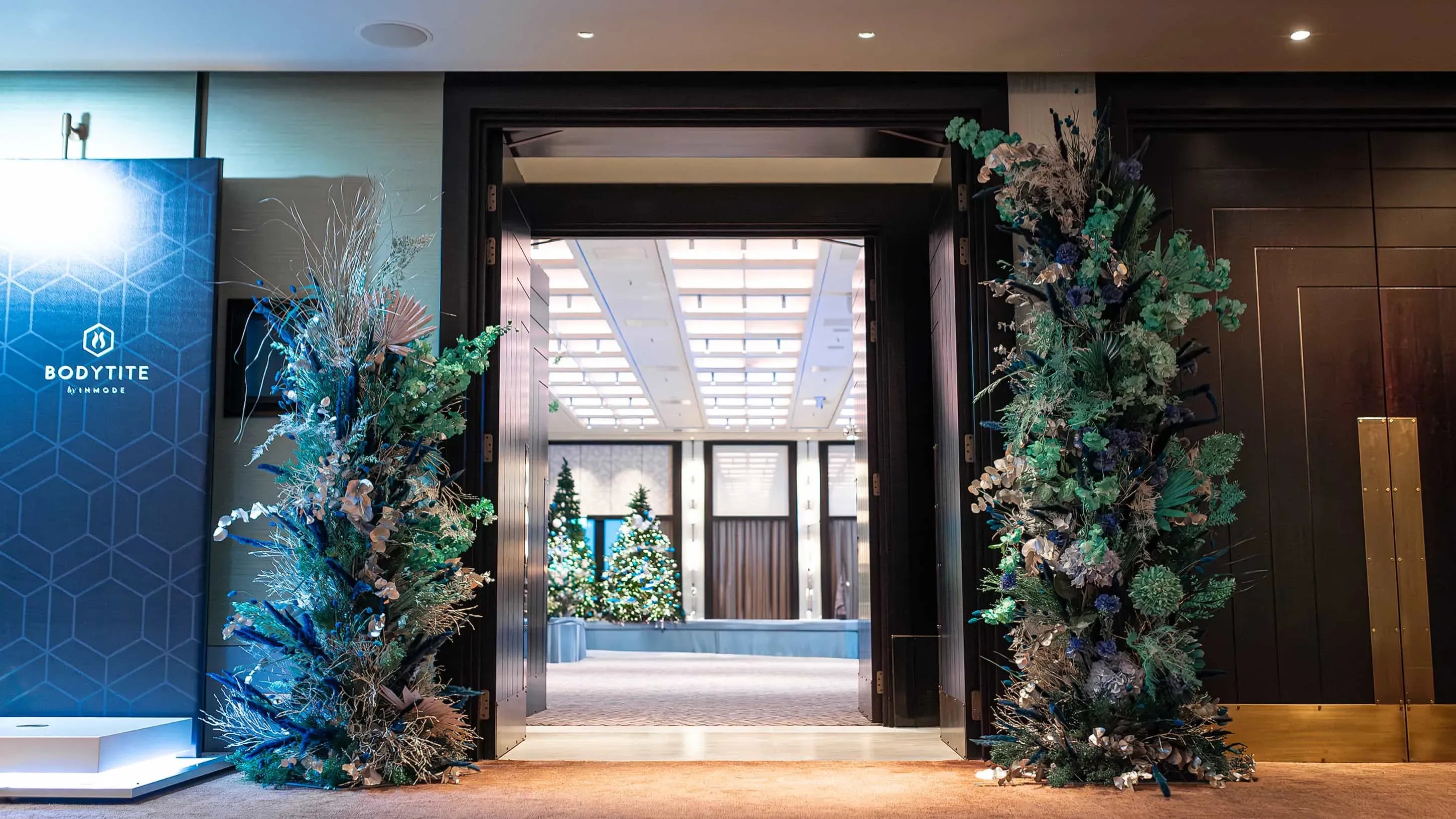 The INMODE welcoming foyer adorned with bespoke festive botanical displays in blue and silver next to a 'BODYTITE by InMode' promotional banner, with a glimpse of a lit Christmas tree through the doorway.