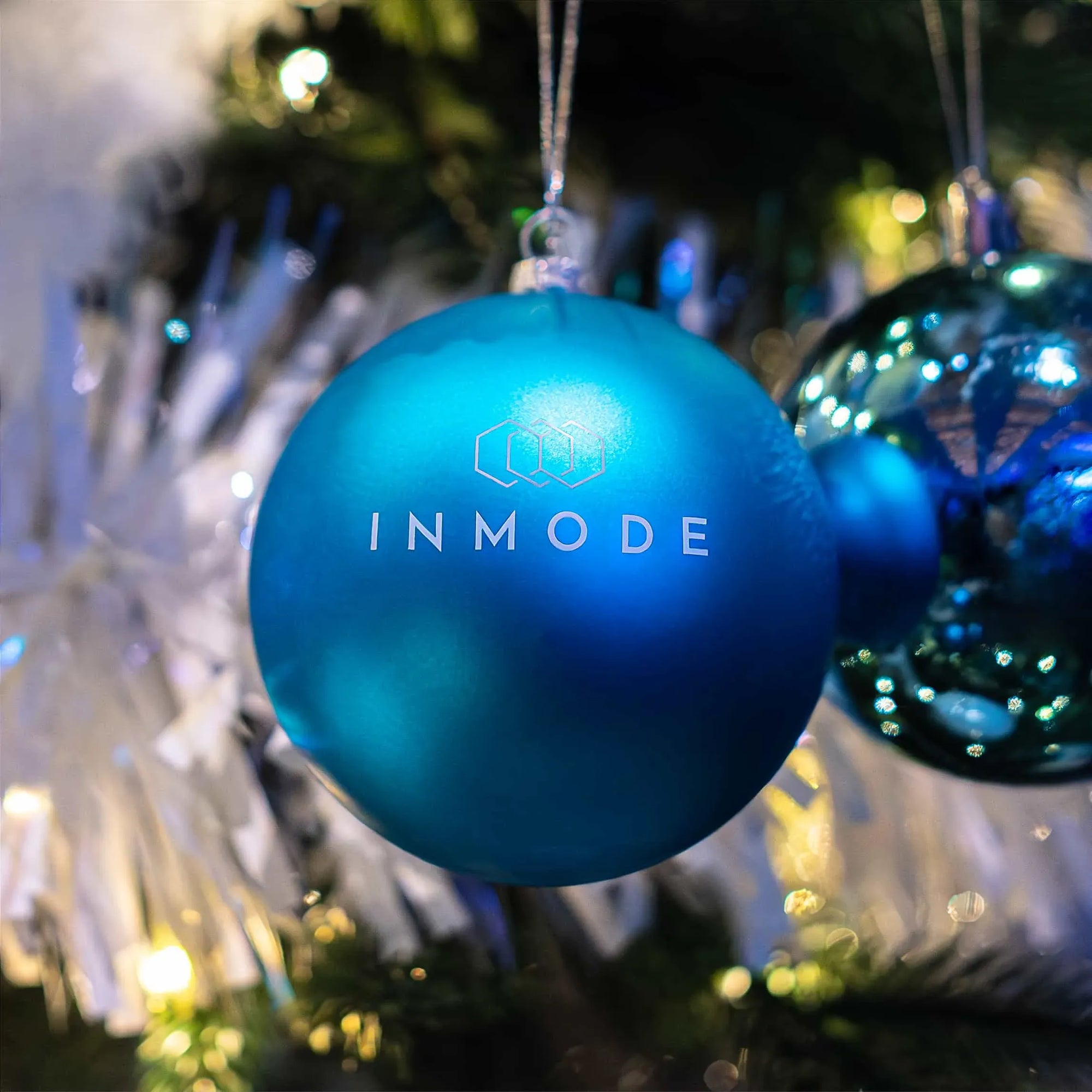 A close-up of a matte blue Christmas bauble with the white 'INMODE' logo on it, set against a backdrop of festive lights and white Christmas tree branches.