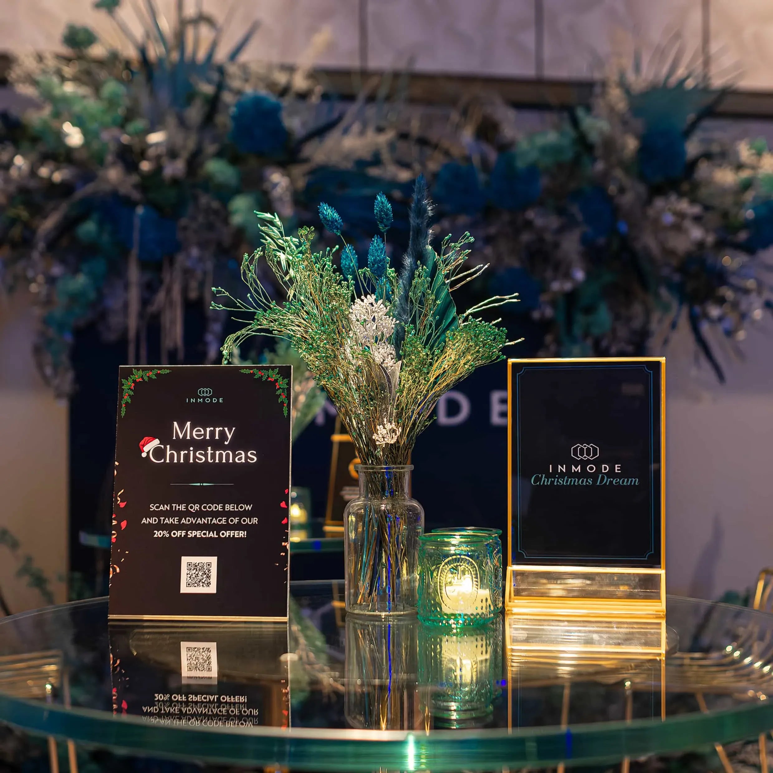An elegant Christmas marketing setup on a glass table, displaying an INMODE 'Merry Christmas' card with a QR code, next to a branded 'INMODE Christmas Dream' plaque, with a vase of festive greenery and lit candles, all set against a backdrop of luxurious blue floral decorations. designed and created by Amarantè London.