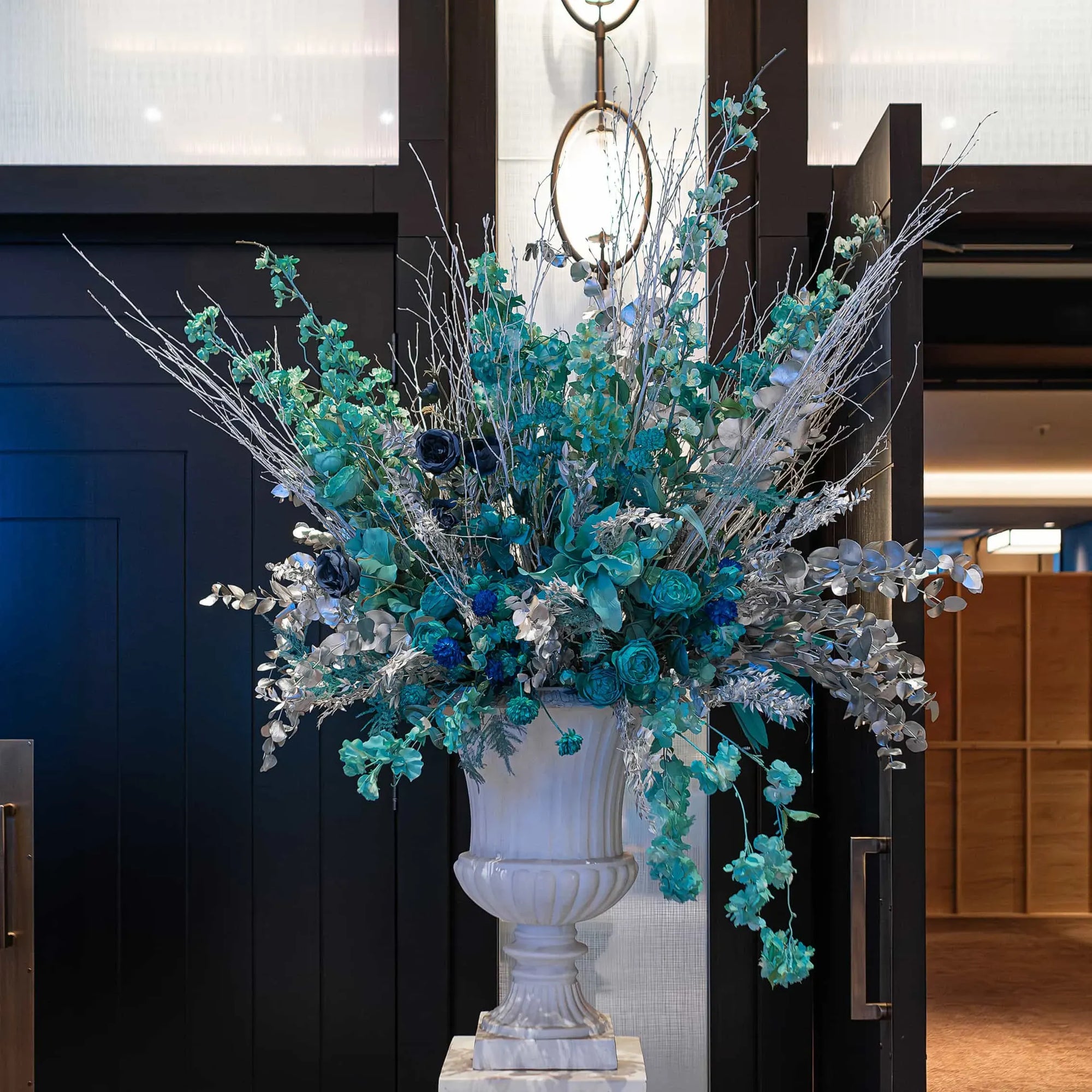 A grand floral display with blue roses, hydrangeas, and silver foliage arranged in an ornate white urn, set against a dark paneled wall and a modern circular light fixture in the background.
