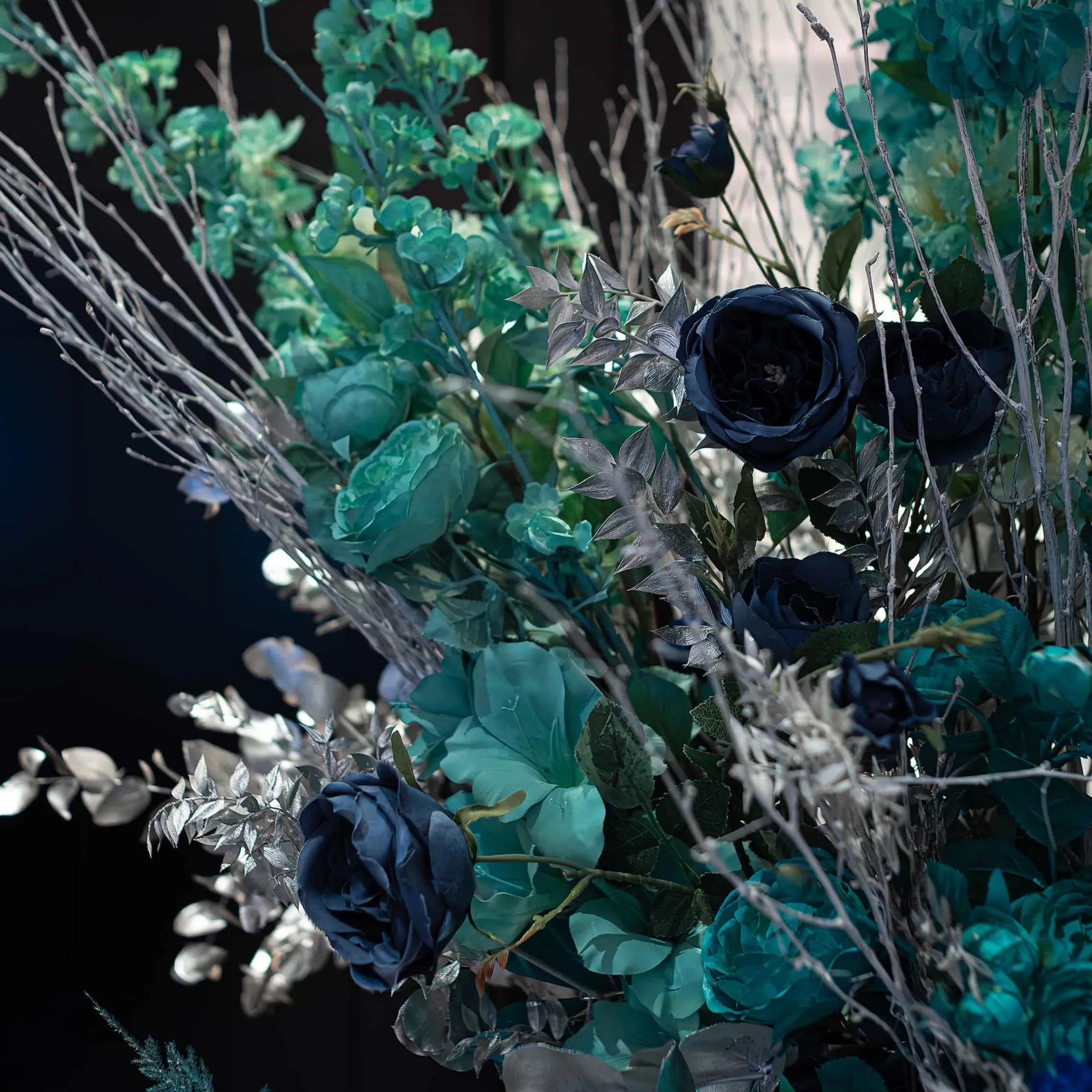 Close-up of a lush arrangement featuring deep navy blue roses, teal hydrangeas, and silver leaves, with delicate branches adding texture against a dark background.