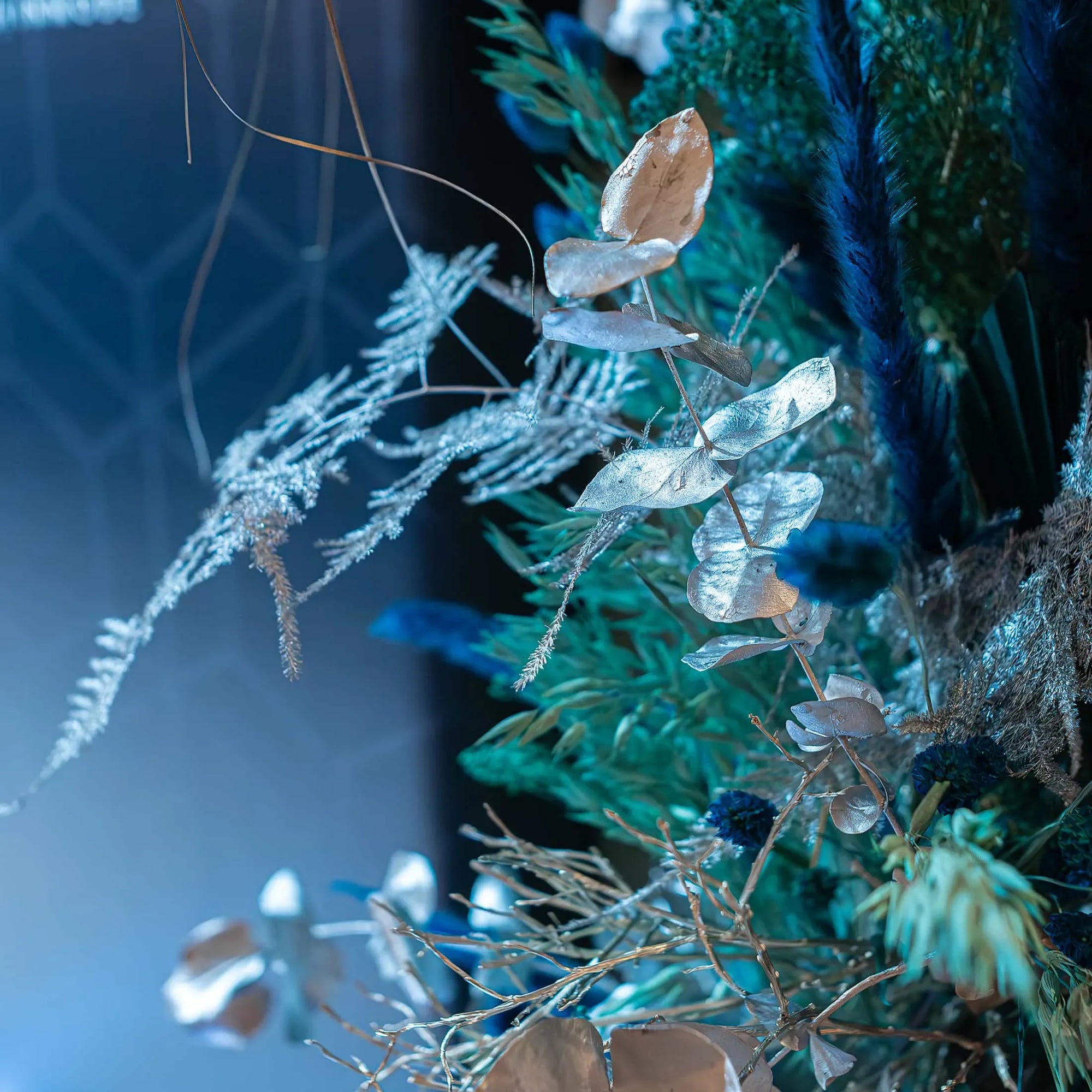 Close-up of bespoke Christmas floral arrangement by Amaranté London for the InMode event at Nobu Hotel. Features silver eucalyptus leaves, ferns, and blue feathers, creating a festive and luxurious atmosphere with a silver and blue colour palette.