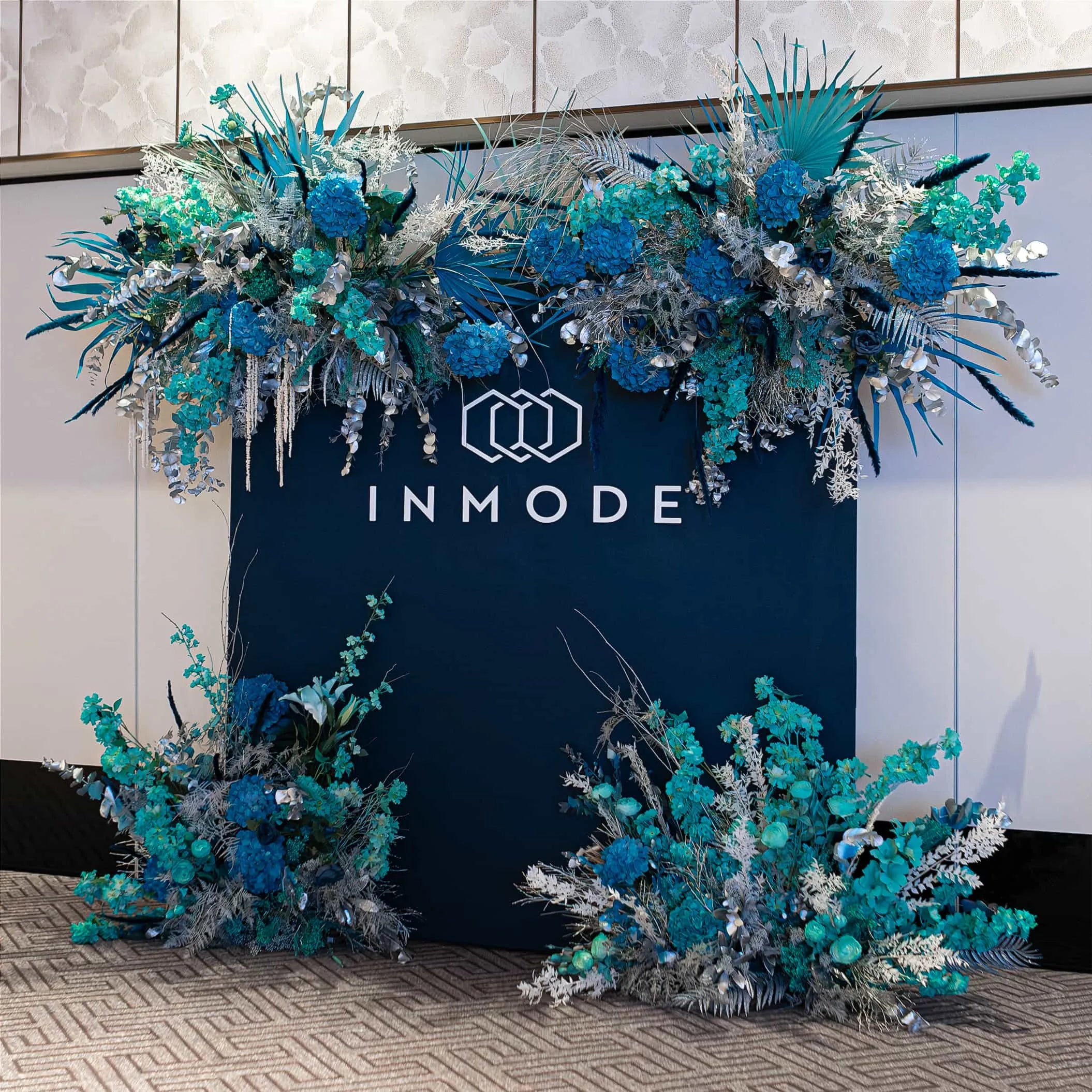 Elegant floral arrangement in blue, teal, and silver hues crowning a dark backdrop with the INMODE logo, creating a festive atmosphere in a contemporary setting with textured wall panels.