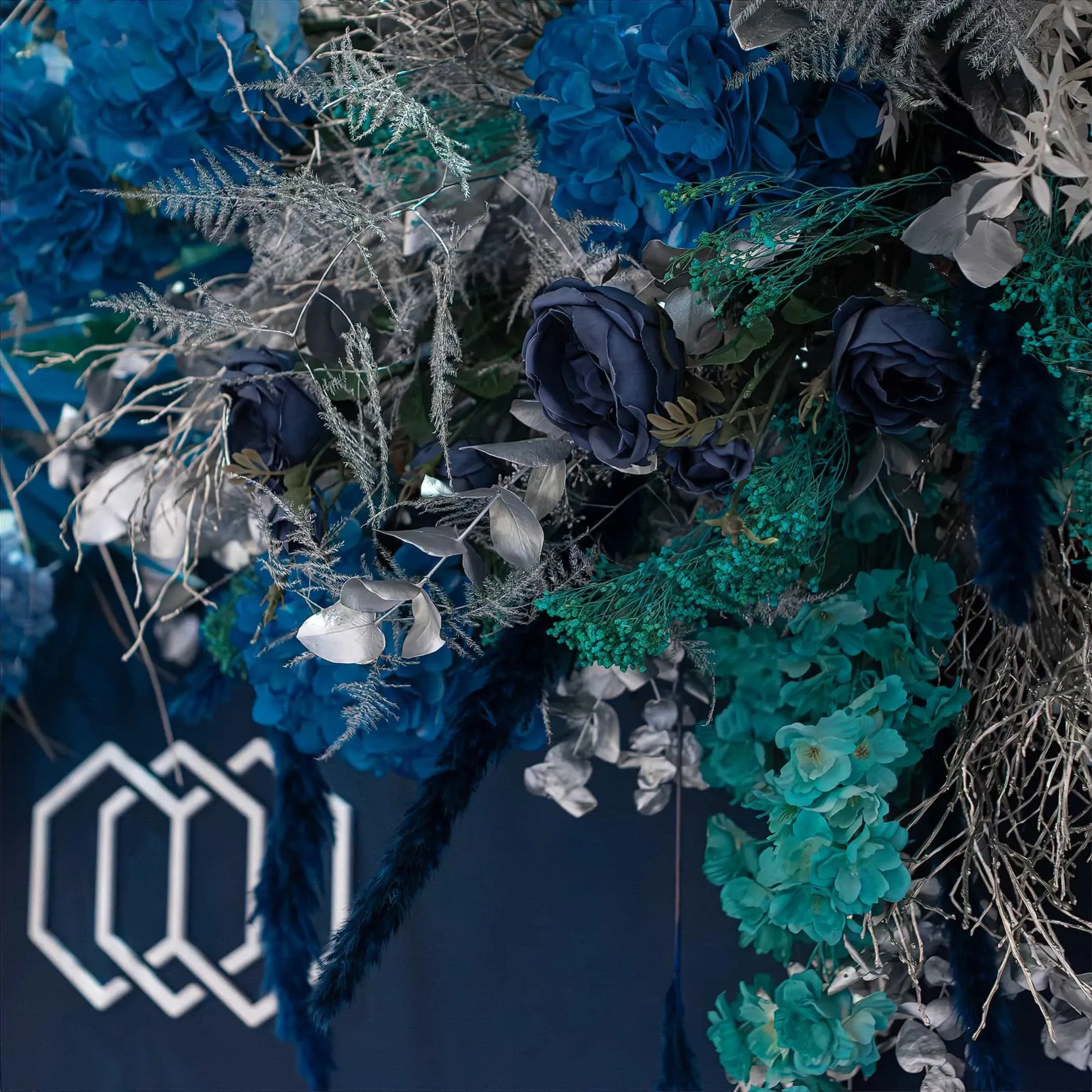 A close-up of a sophisticated botanical arrangement for Christmas with navy blue floral accents, soft blue-green hydrangeas, and silvery leaves, set before a backdrop with a stylized white hexagonal INMODE logo.