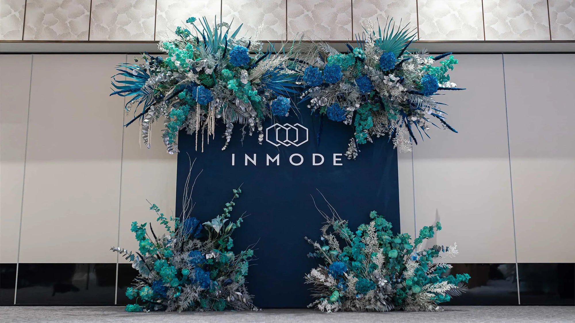 An Elegant Christmas floral arrangement in blue, teal, and silver hues crowns a dark backdrop with the INMODE logo, creating a festive atmosphere in a contemporary setting with textured wall panels, designed, developed, and installed by Event Florist Amaranté London.
