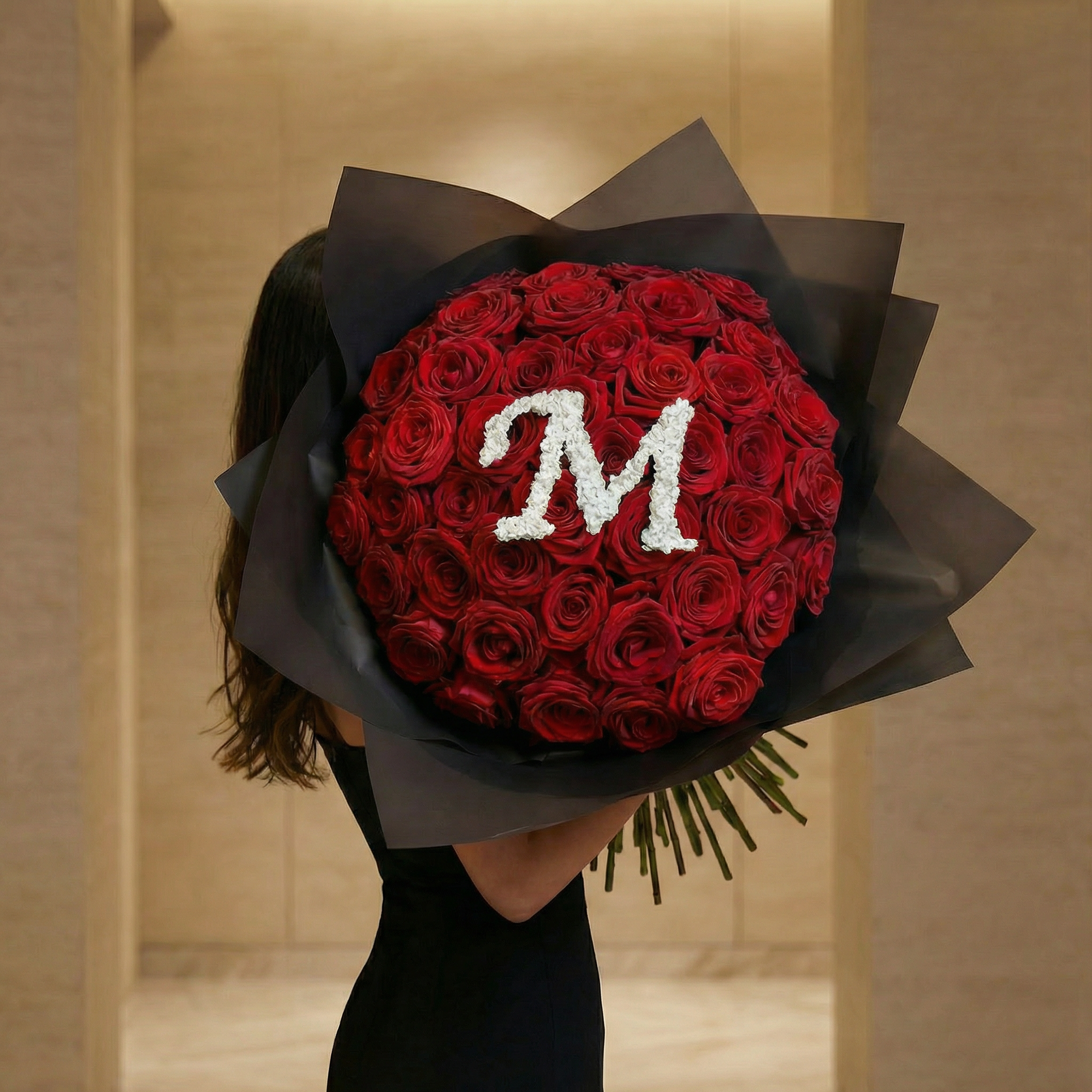 Woman holding exquisite red rose bouquet featuring white gypsophila letter 'M' design, wrapped in premium charcoal paper with dramatic presentation in warm interior setting - Amarante London