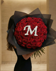 Woman holding exquisite red rose bouquet featuring white gypsophila letter 'M' design, wrapped in premium charcoal paper with dramatic presentation in warm interior setting - Amarante London