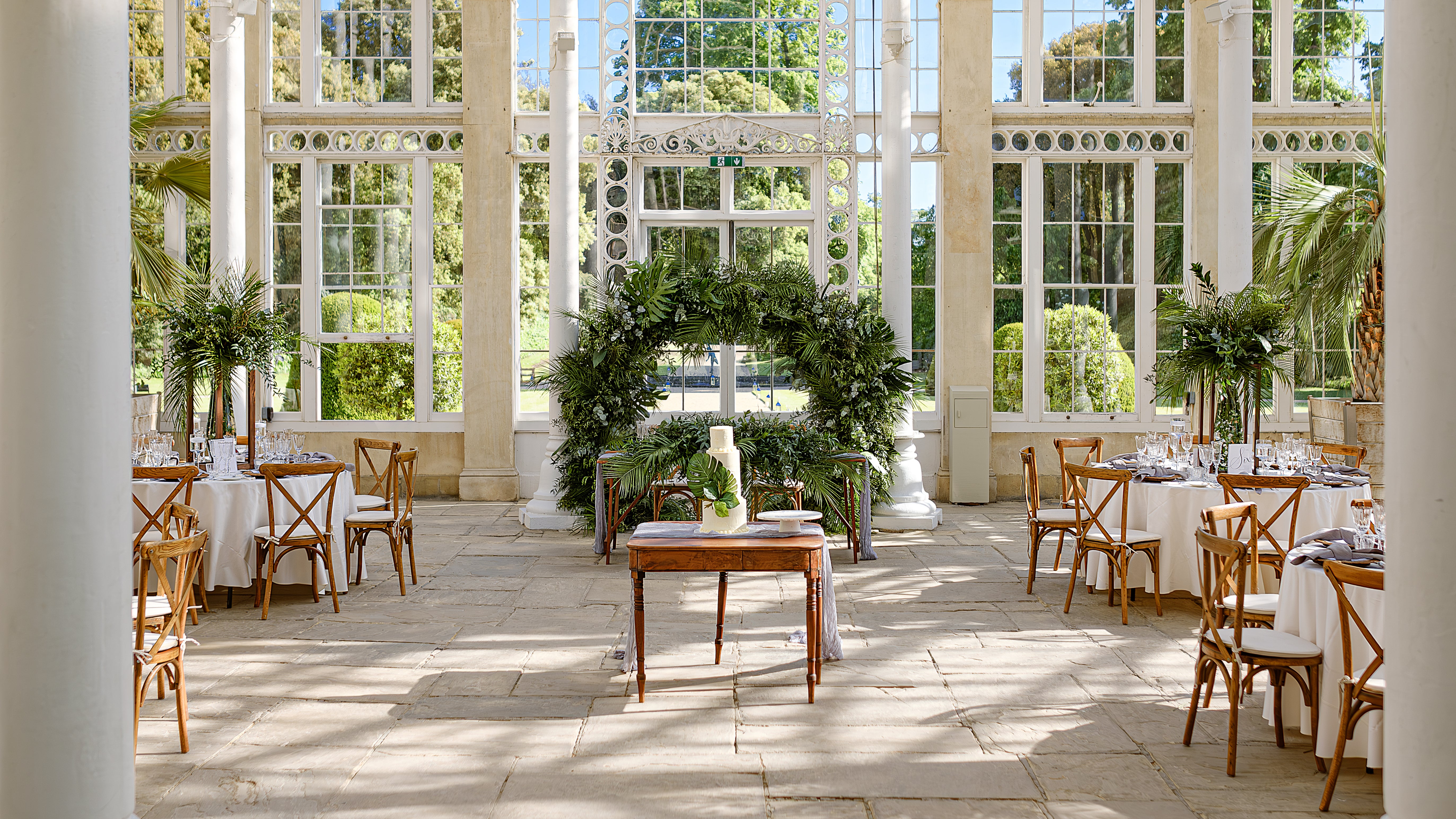 Wedding ceremony at Londo Hilton Syon Park, conservatory with large foliage wreath, wooden cross-back chairs, and floor-to-ceiling windows overlooking gardens - Floral Design by Amaranté London.