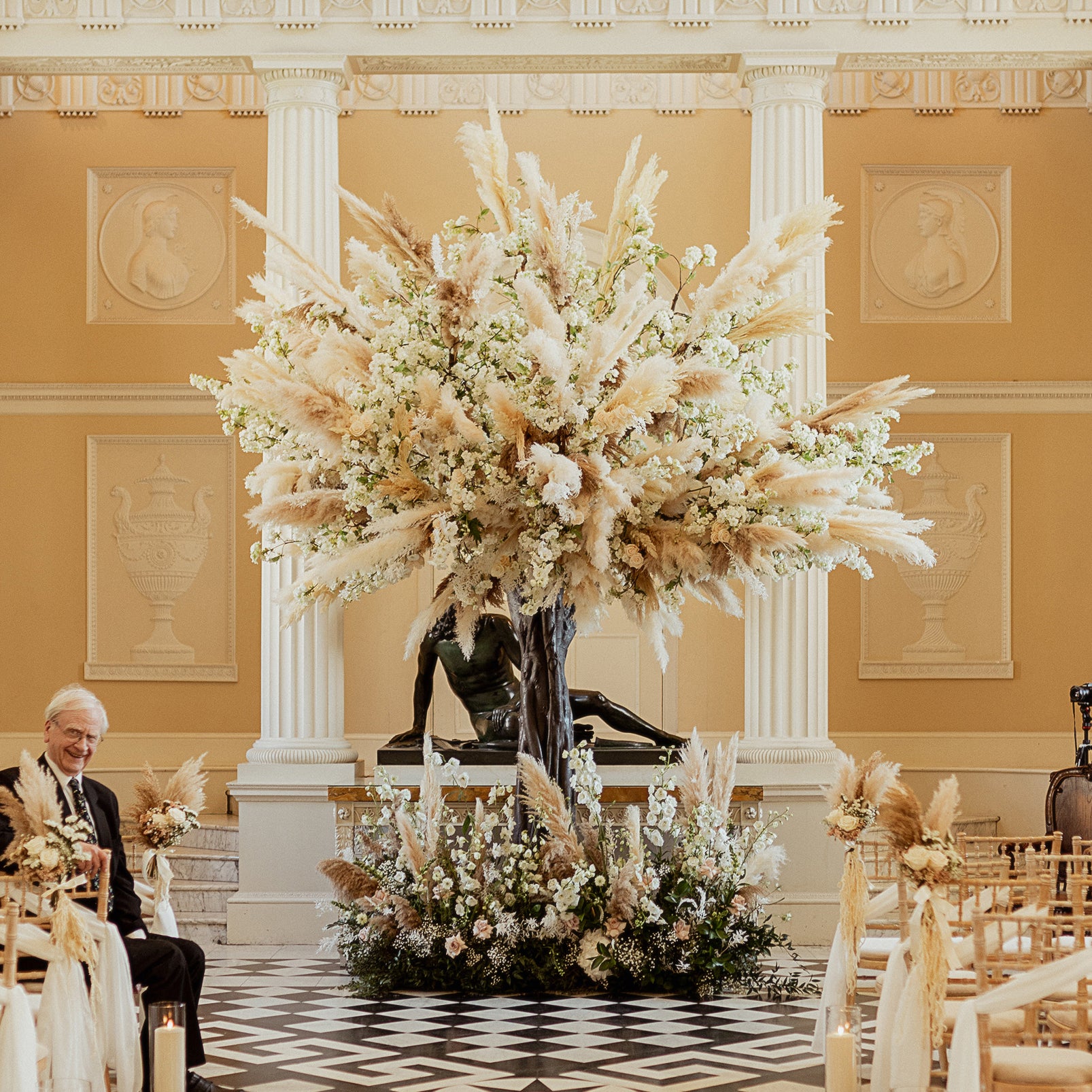 Wedding ceremony centerpiece at London Holton Syon Park with oversized pampas grass and white floral arrangement in neoclassical Great Hall with checkerboard floor - Floral Design by Amaranté London