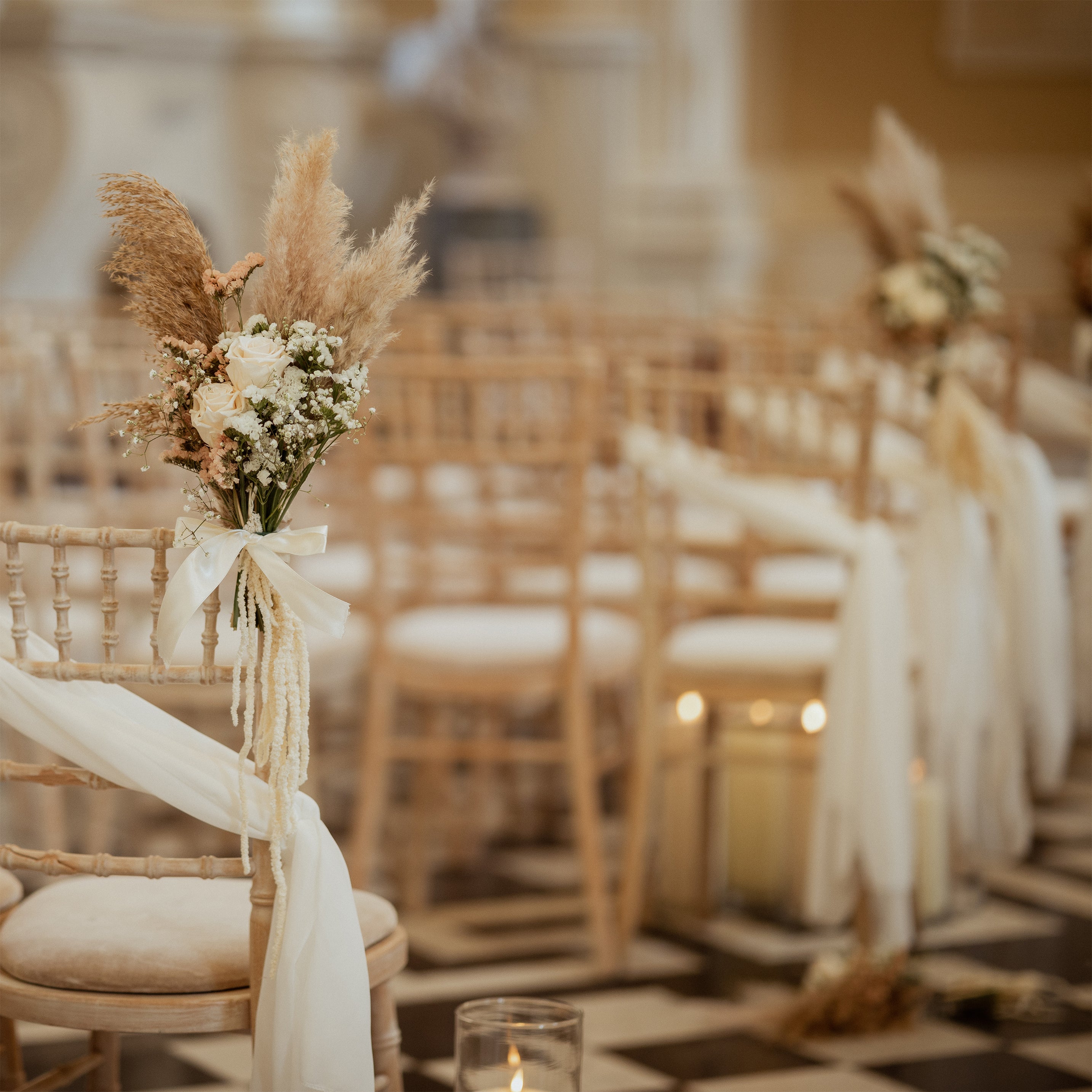 Wedding ceremony aisle at London Hilton Syon Park with pampas grass and white rose arrangements tied to gold chiavari chairs with ivory ribbon - Floral Design by Amaranté London