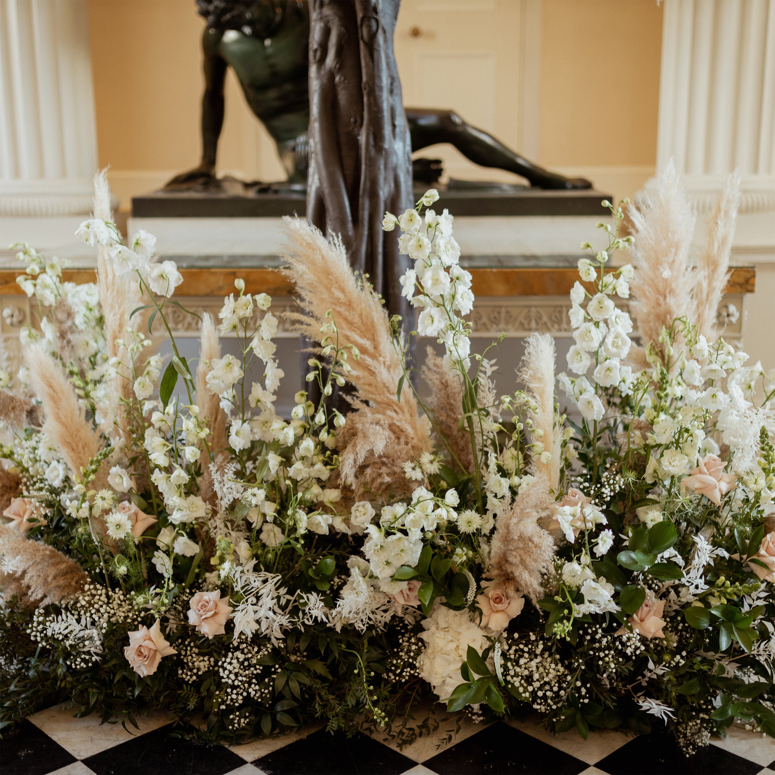 Luxurious floral arrangement at Syon Park with white delphiniums, blush roses, pampas grass, and baby's breath against a classical sculpture backdrop - Floral Design by Amaranté London