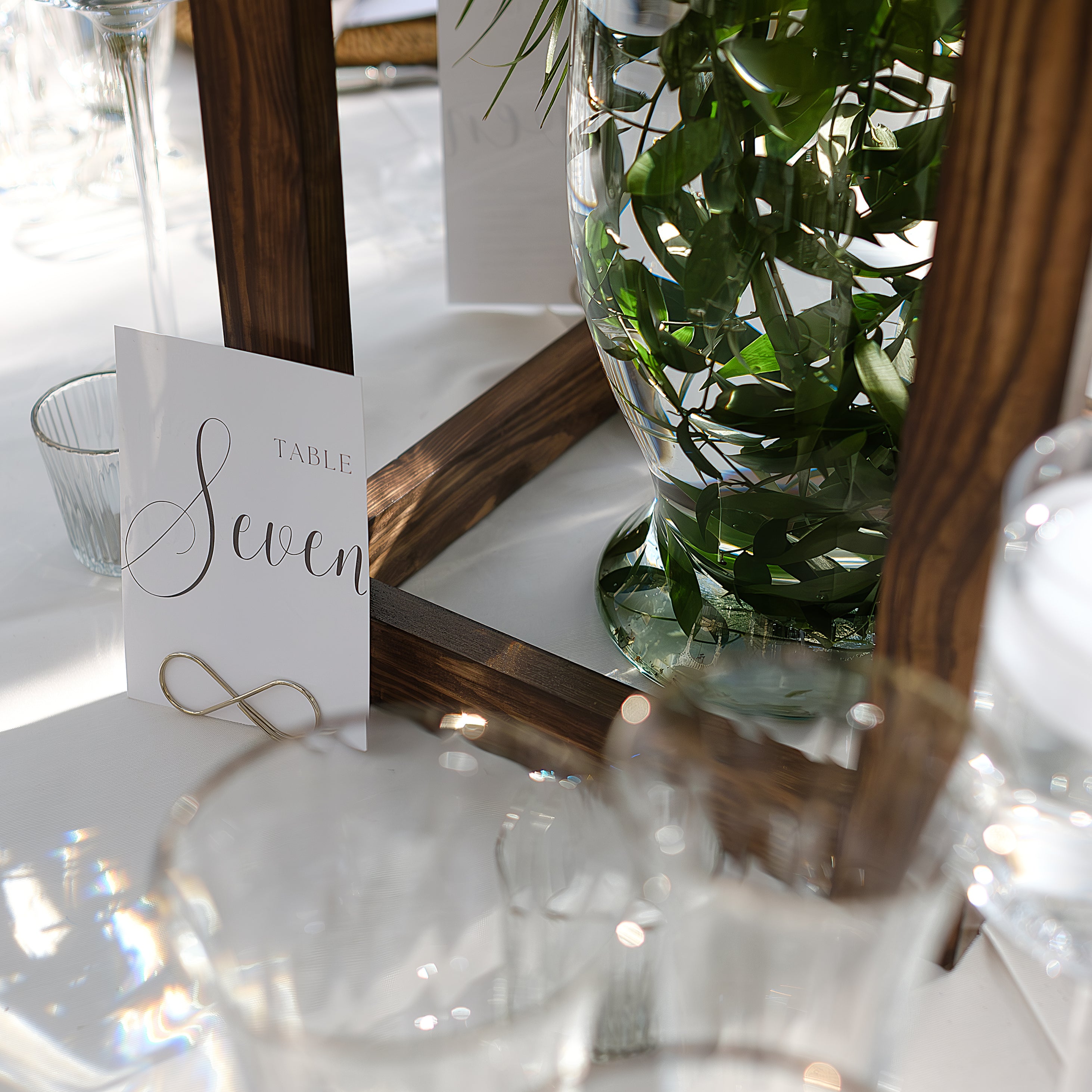 Simple green eucalyptus stems in a clear glass vase beside the table seven card at the London Hilton Syon Park wedding reception - Floral Design by Amaranté London