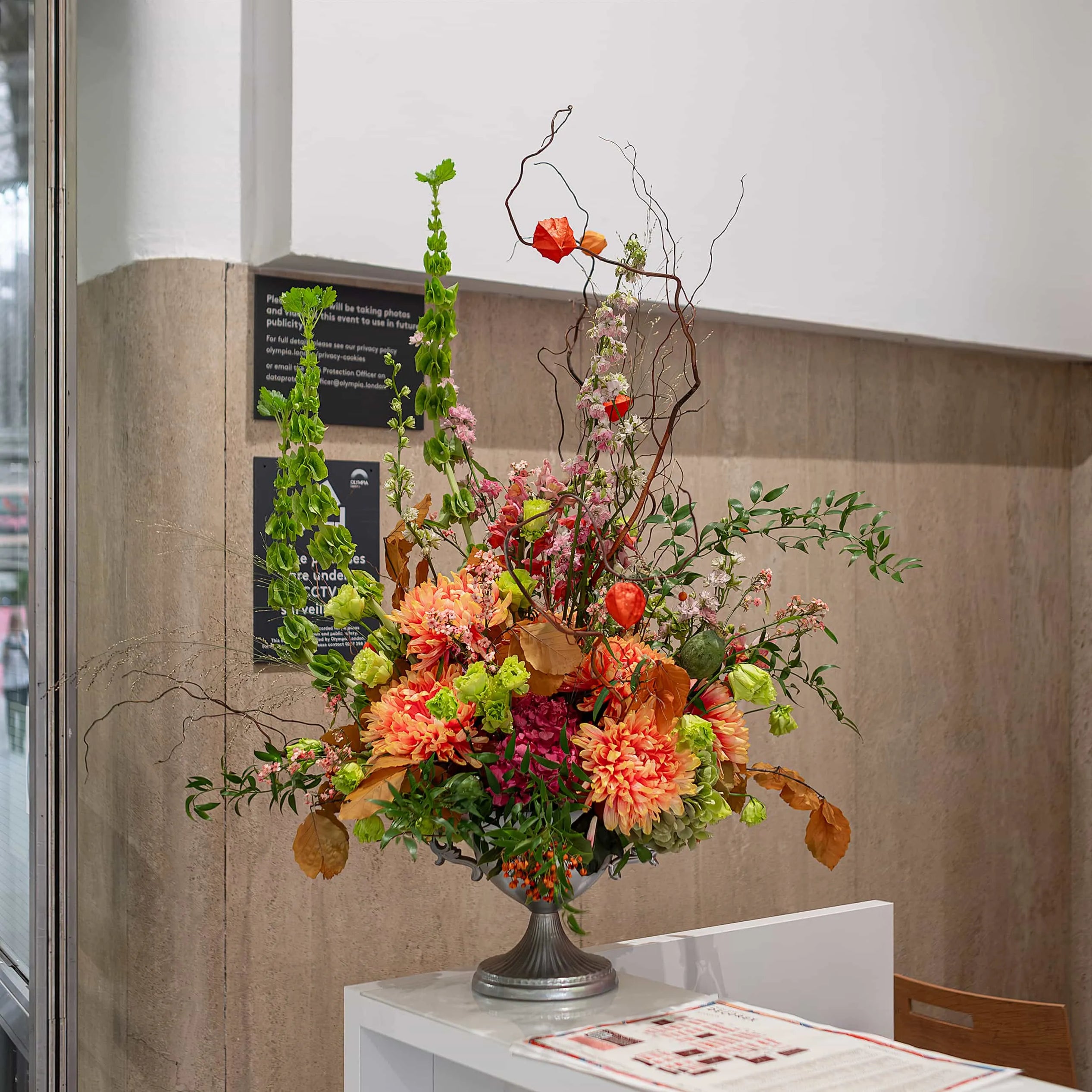 Elegant floral arrangement at Decorex’s registration desk, featuring autumnal tones and lush foliage, combining aesthetics with a warm welcome -by Amaranté London Event Florist.