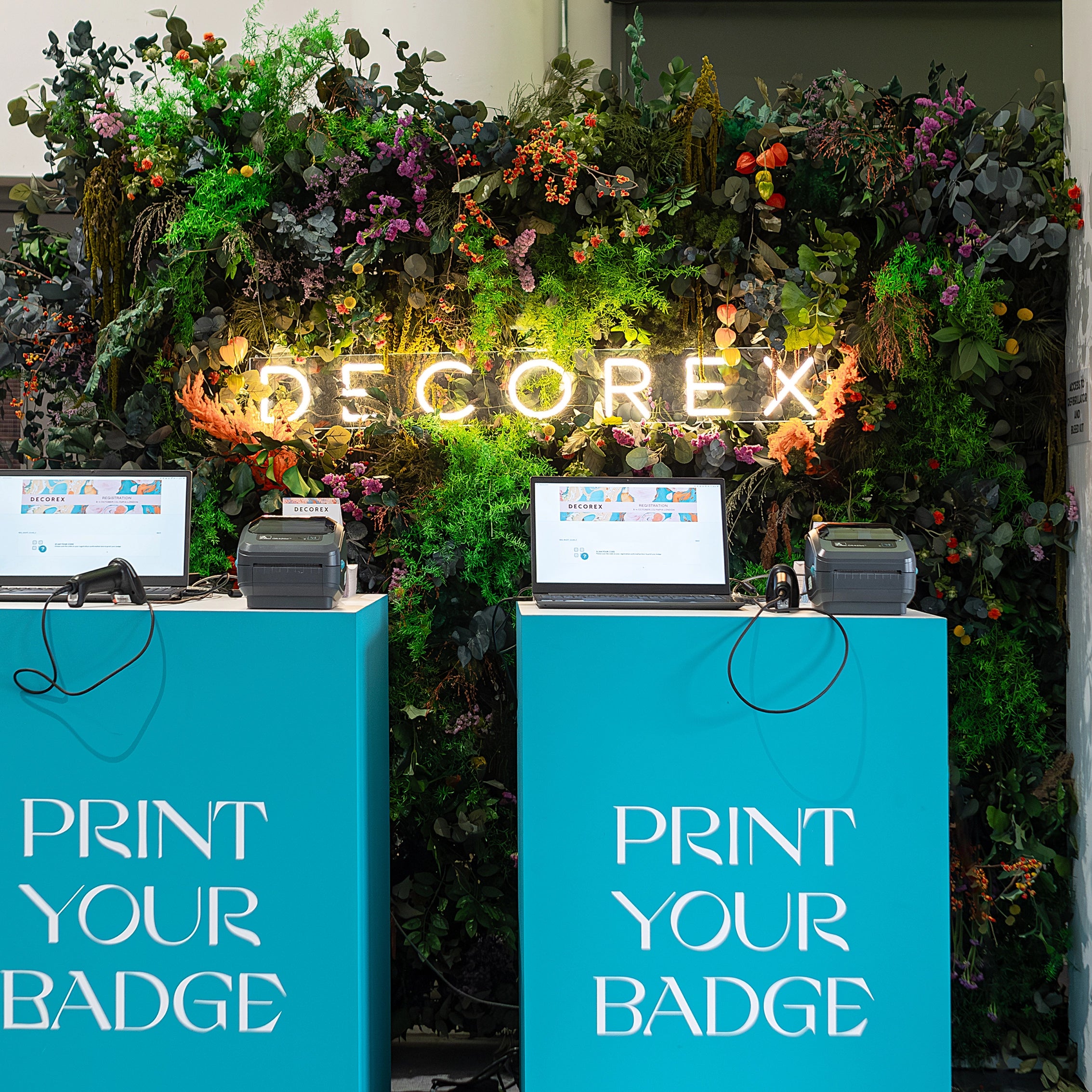 An immersive floral wall installation at Decorex featuring the event’s branding illuminated amidst a cascade of colourful flowers and greenery - Amaranté London Event Florist