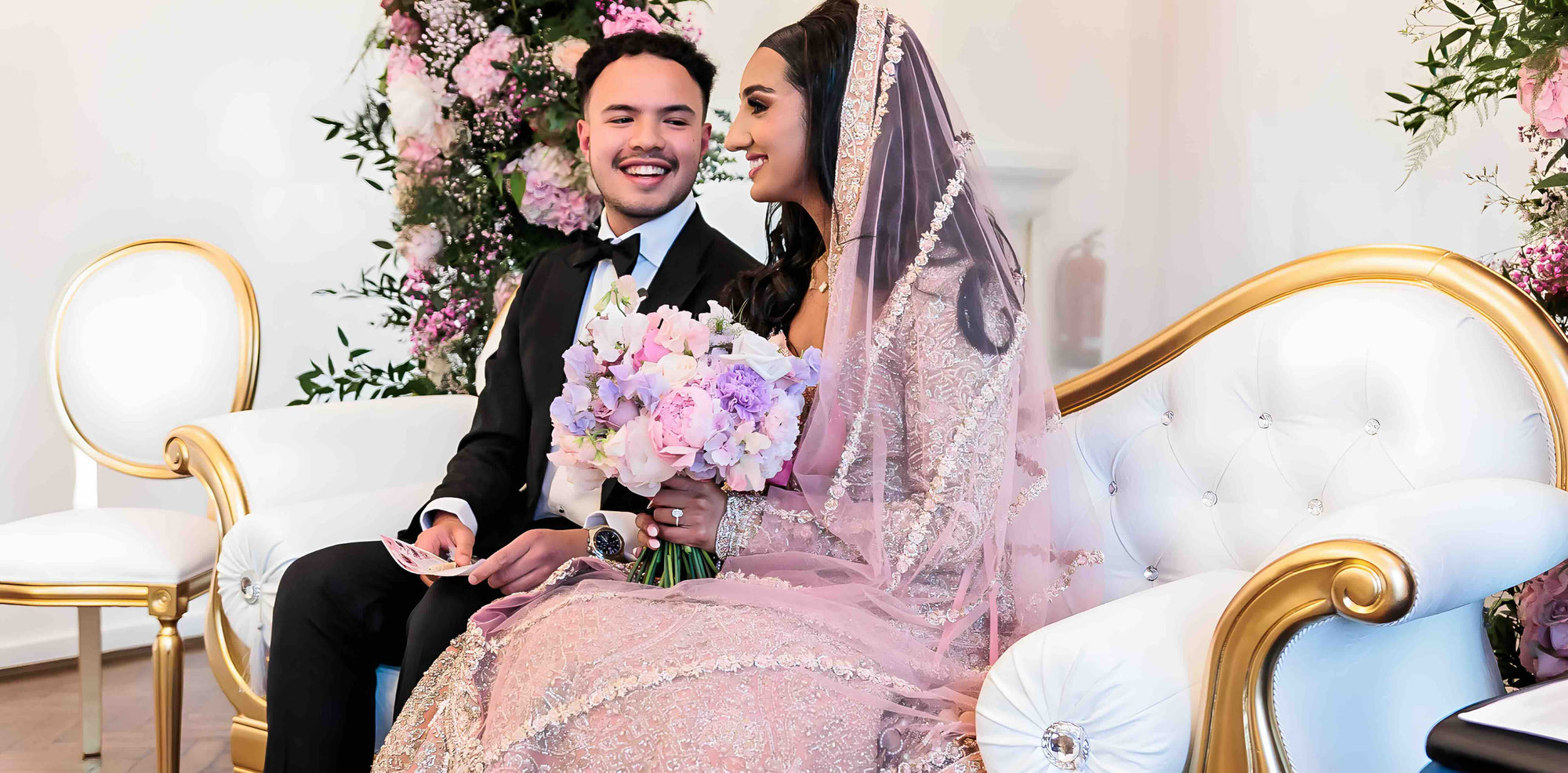 Happy couple on wedding day with pastel bouquet and floral backdrop by Amaranté London wedding planning team