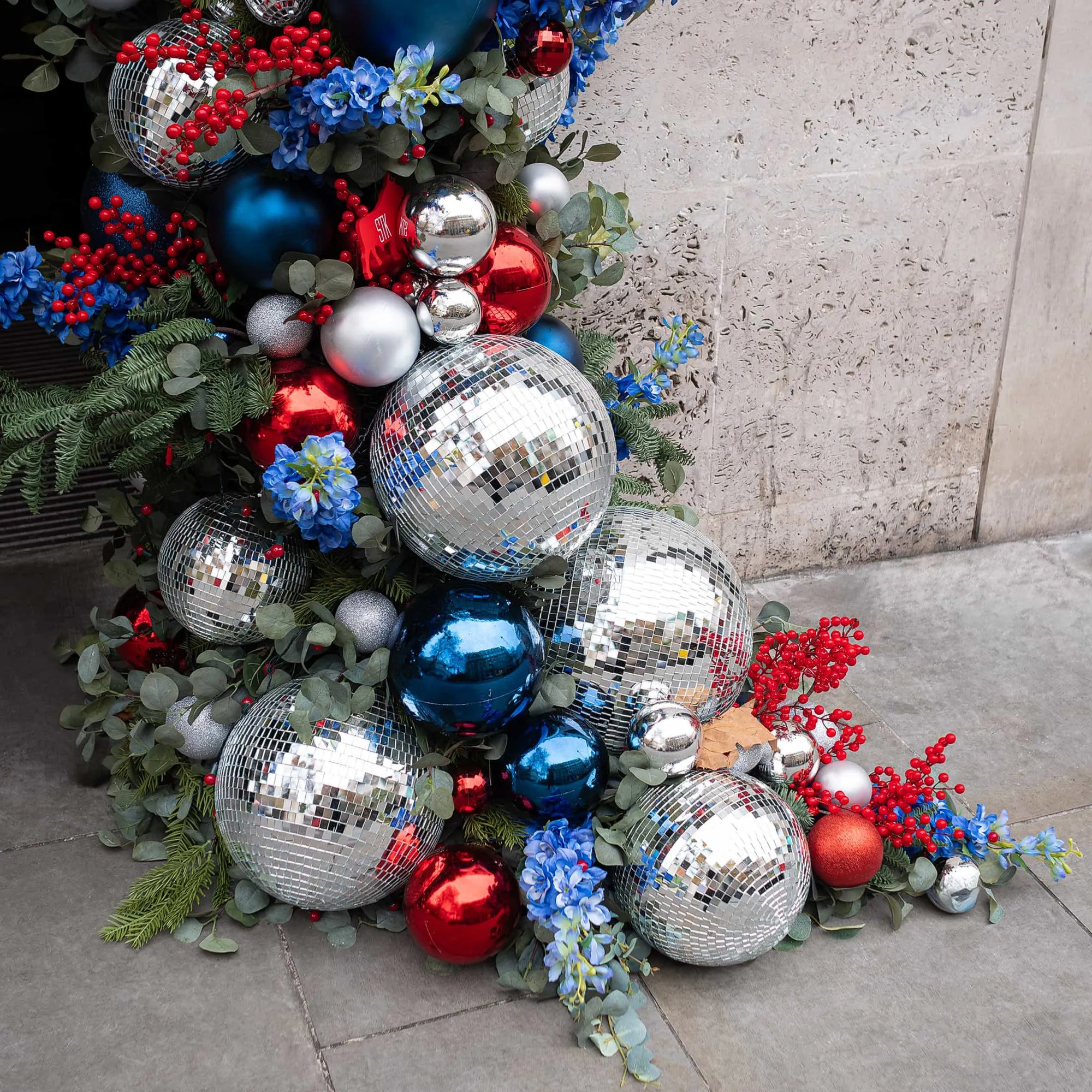 Close-up of the Christmas floral arrangement designed and created by Amaranté London for the STK Steakhouse entrance, featuring a cluster of mirror disco balls, red and blue baubles interspersed with blue hydrangea flowers and red holly berries amidst evergreen foliage.