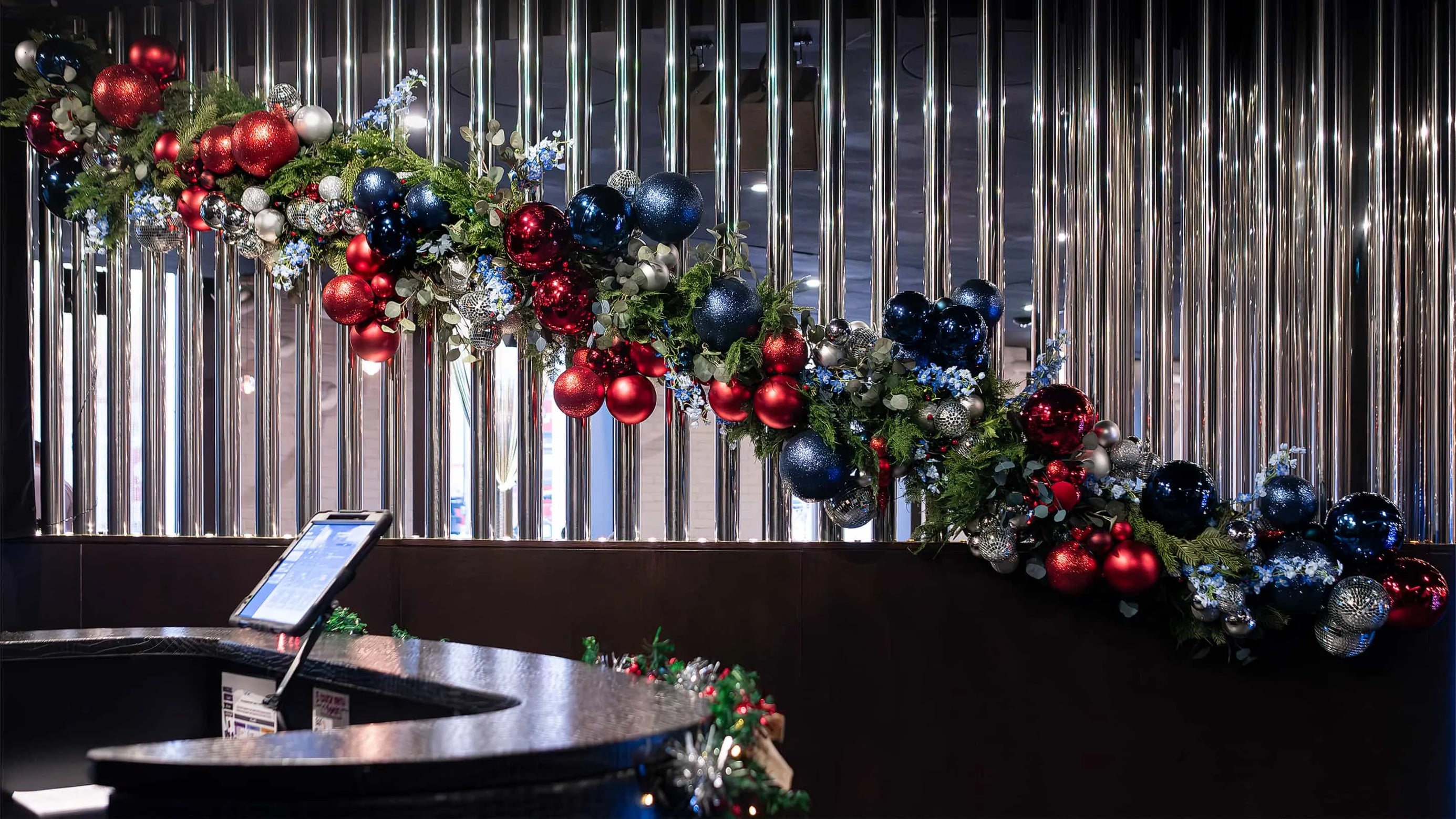 Luxuriously decorated garland with red and blue Christmas baubles and silver accents draping over the reception counter at STK Steakhouse, complemented by a backdrop of reflective vertical metal strips.