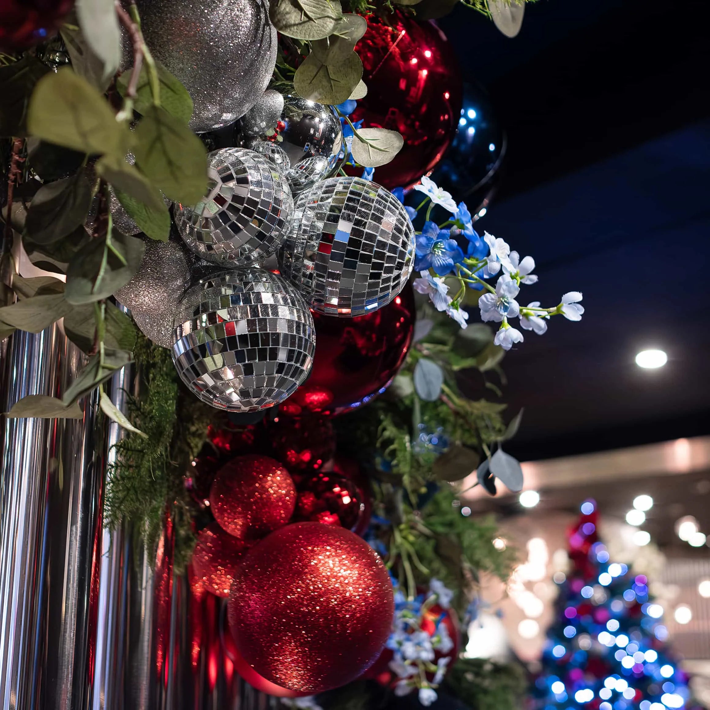 Side view of a Christmas garland on a shiny metal backdrop at STK Steakhouse, featuring glittering disco balls, red baubles, and delicate blue flowers amidst greenery, with a Christmas tree in the background.