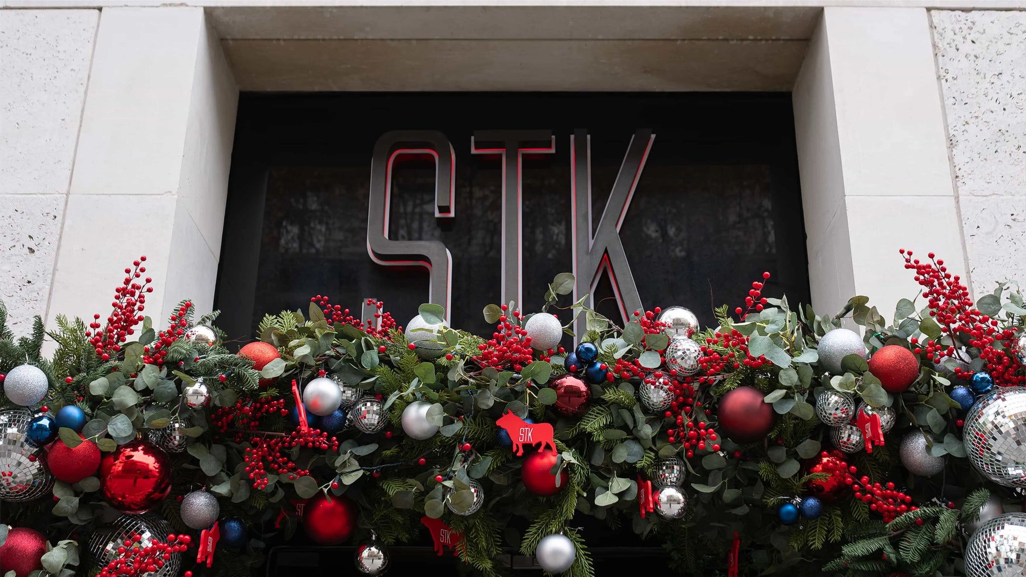 A large luxury external Christmas floral arrangement above the entrance of STK Steakhouse in London with an assortment of red and blue baubles, silver disco balls, and abundant red berry sprigs against a lush backdrop of greenery, designed and created by Event Florist Amarante London.