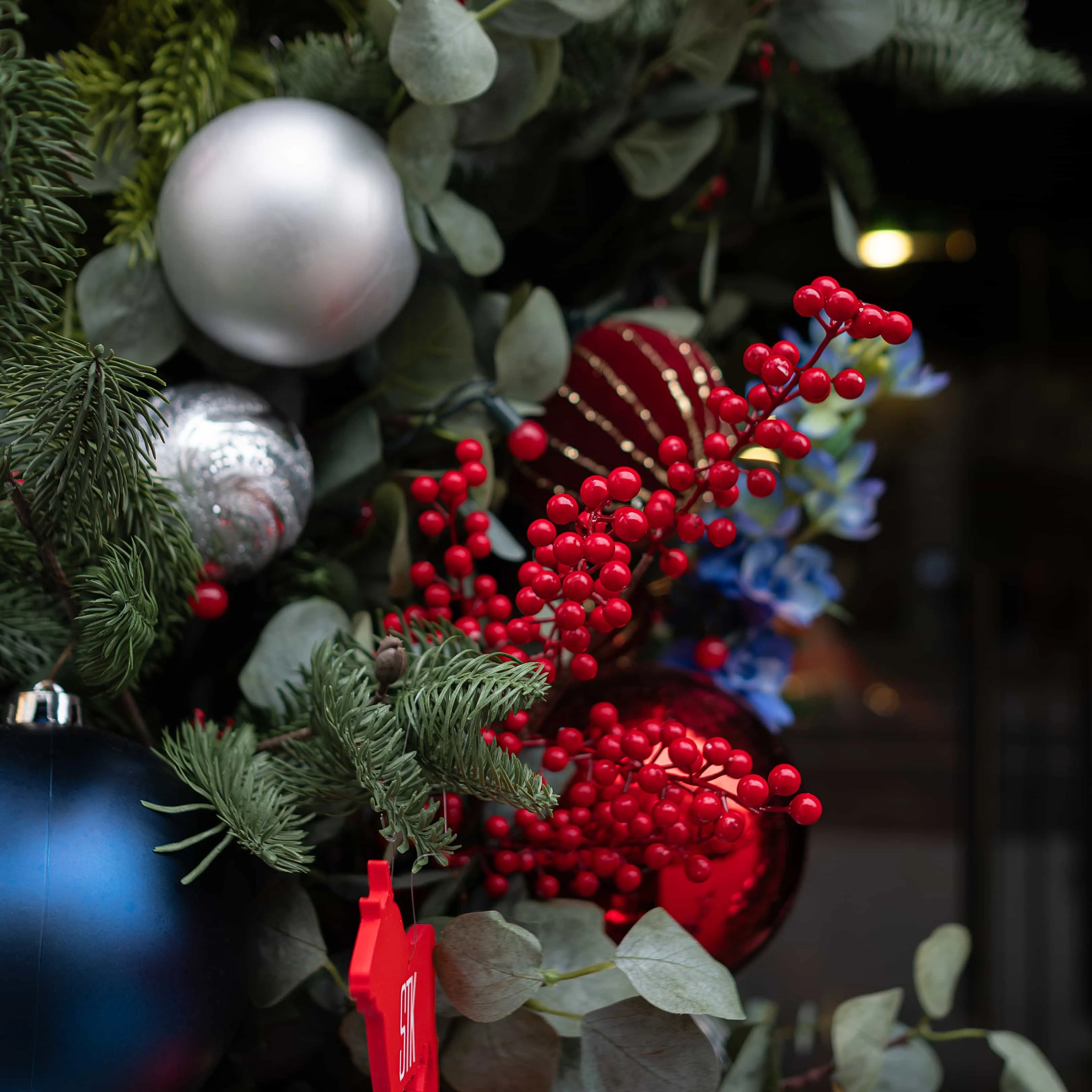 Detailed view of a Christmas floral arrangement at STK Steakhouse, highlighting shiny silver and red ornaments, bright red berry clusters, and a mixture of evergreen and eucalyptus leaves.