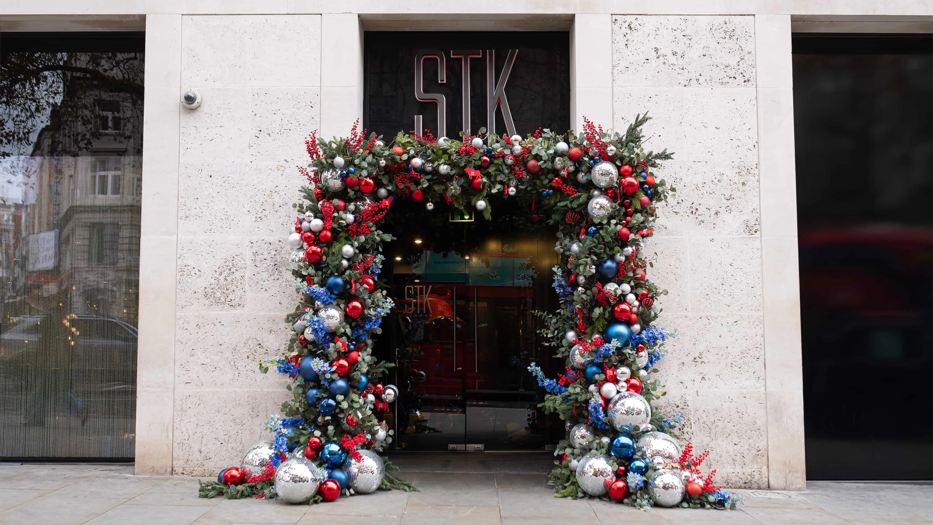Entrance of STK Steakhouse in London, adorned with an elaborate Christmas floral archway, featuring a vibrant mix of red and blue baubles, silver ornaments, and lush greenery.