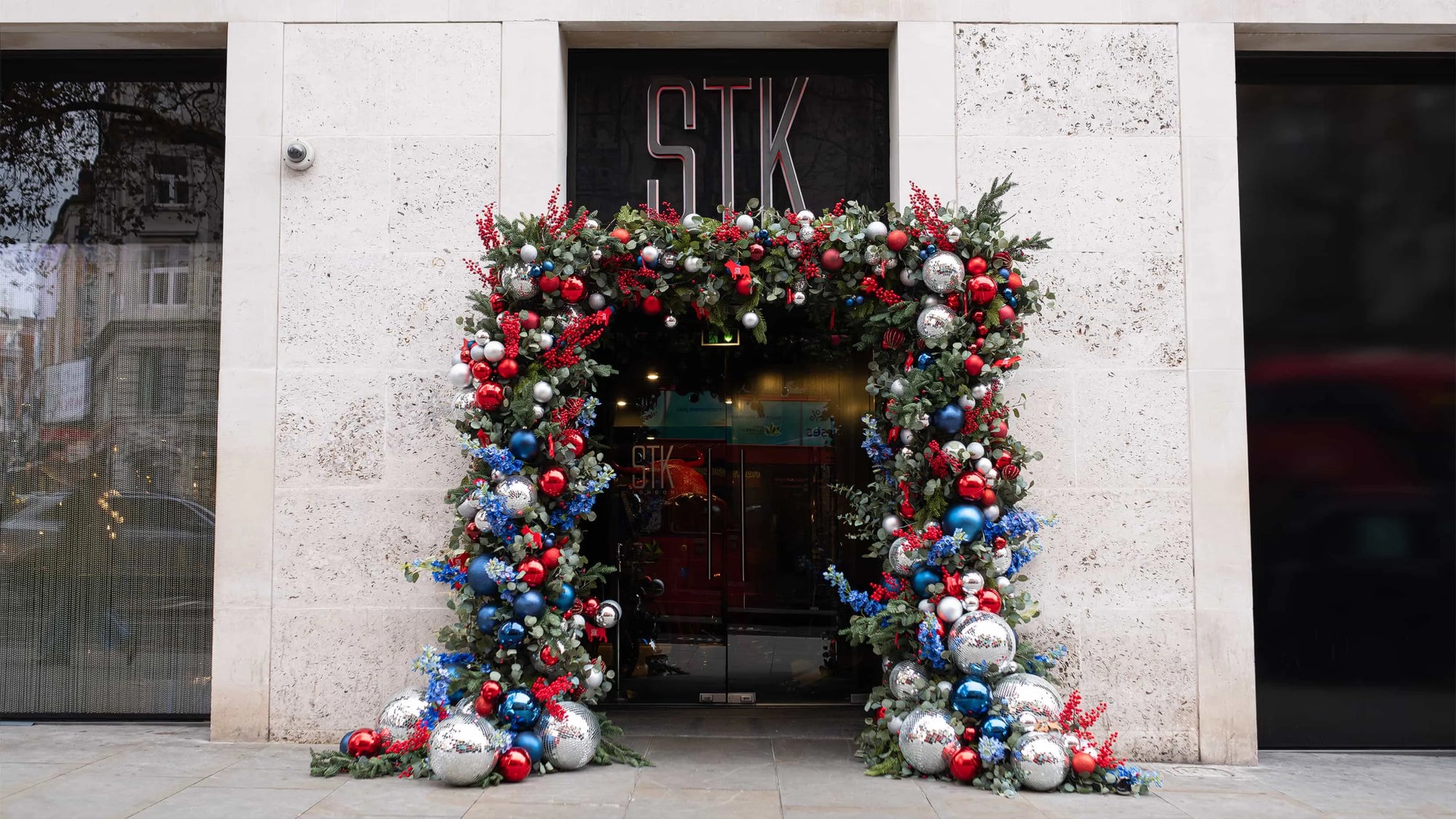 The entrance of STK Steakhouse in London is adorned with an elaborate Christmas floral archway featuring a vibrant mix of red and blue baubles, silver ornaments, and lush greenery.