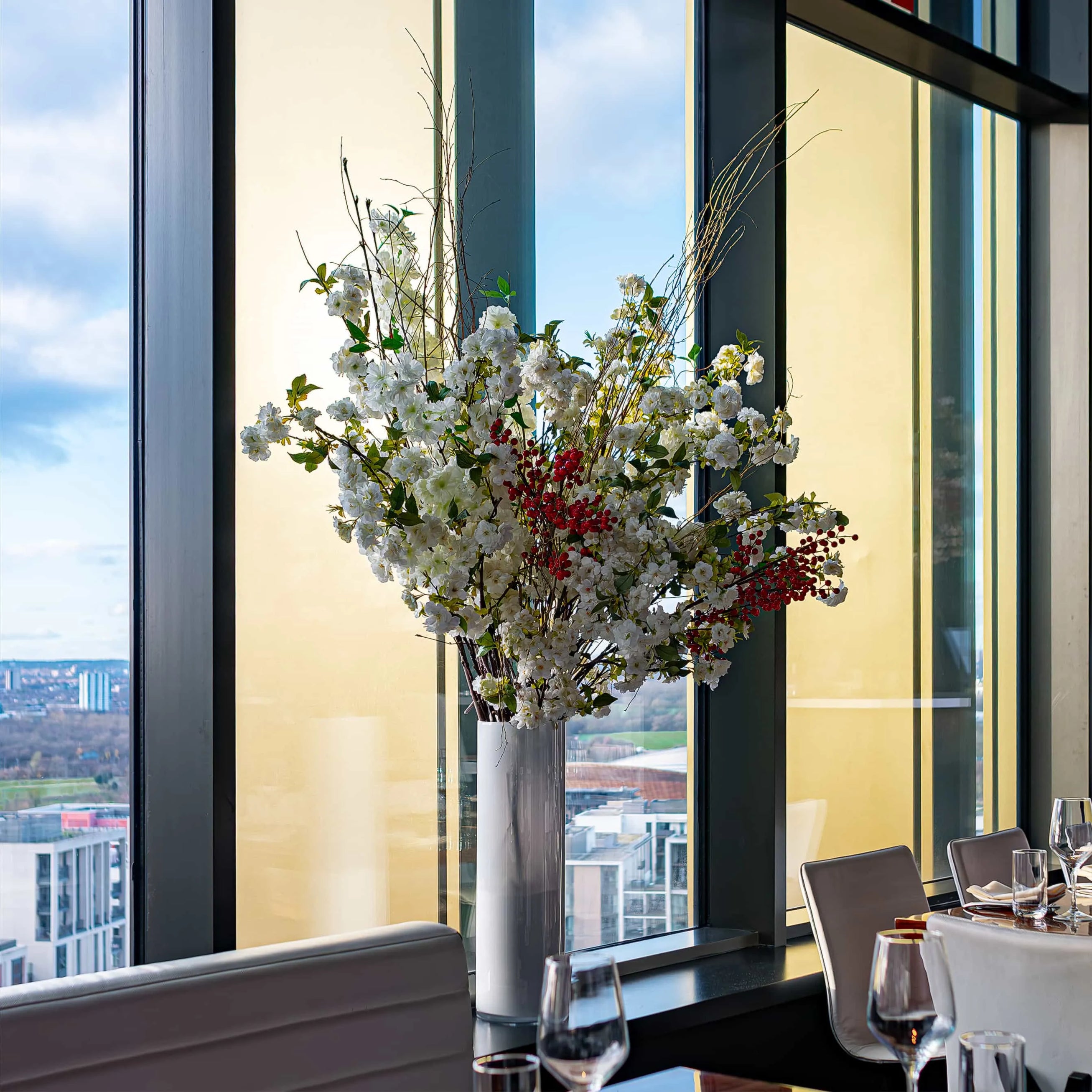 Elegant floral arrangement in a tall vase on a table at STK Steakhouse, with a backdrop of floor-to-ceiling windows offering a view of the urban landscape and blue sky.