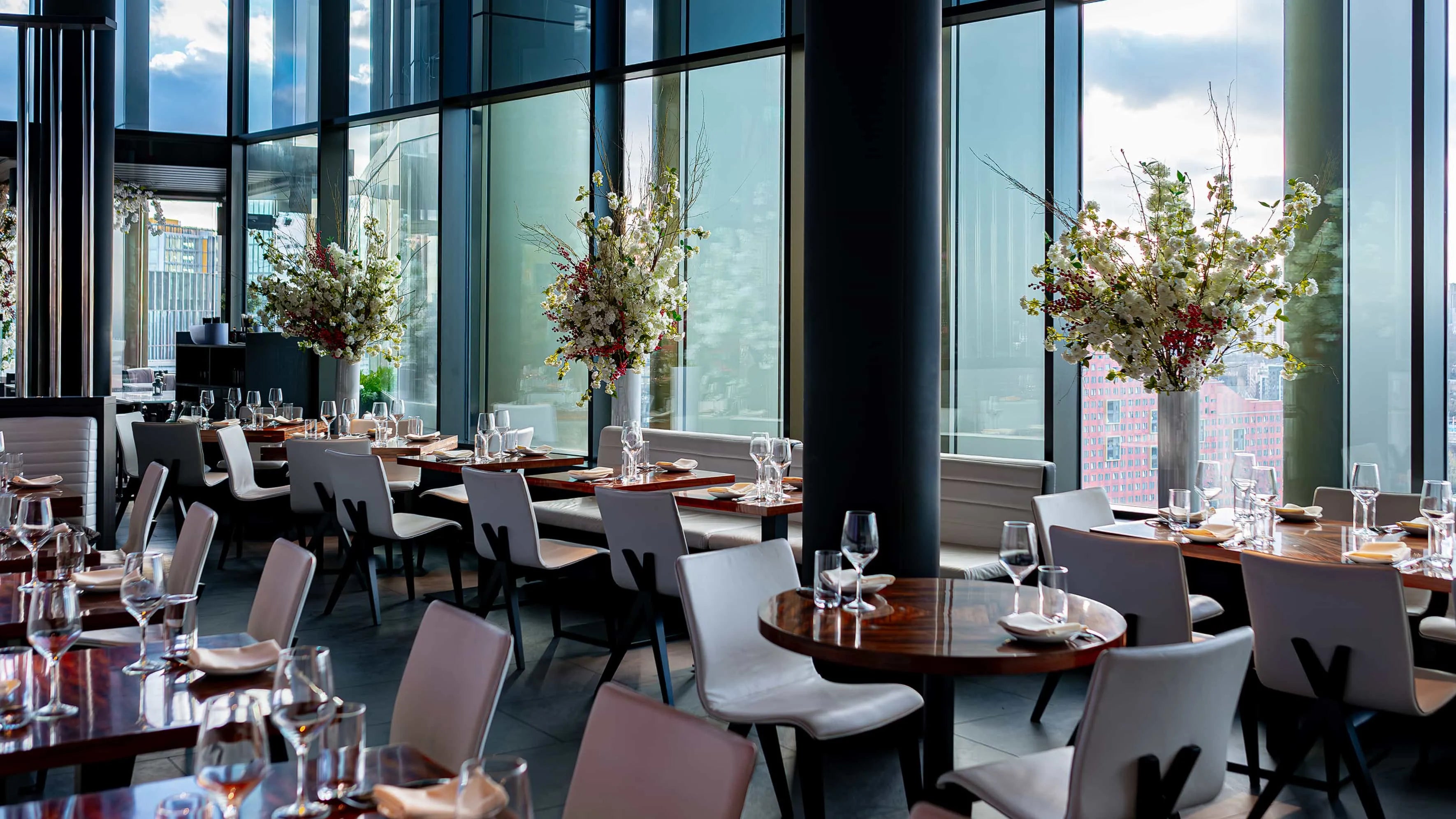 Spacious dining area at STK Steakhouse with elegantly set tables and large floral centrepieces, featuring white blossoms and red accents, beside floor-to-ceiling windows offering city views.