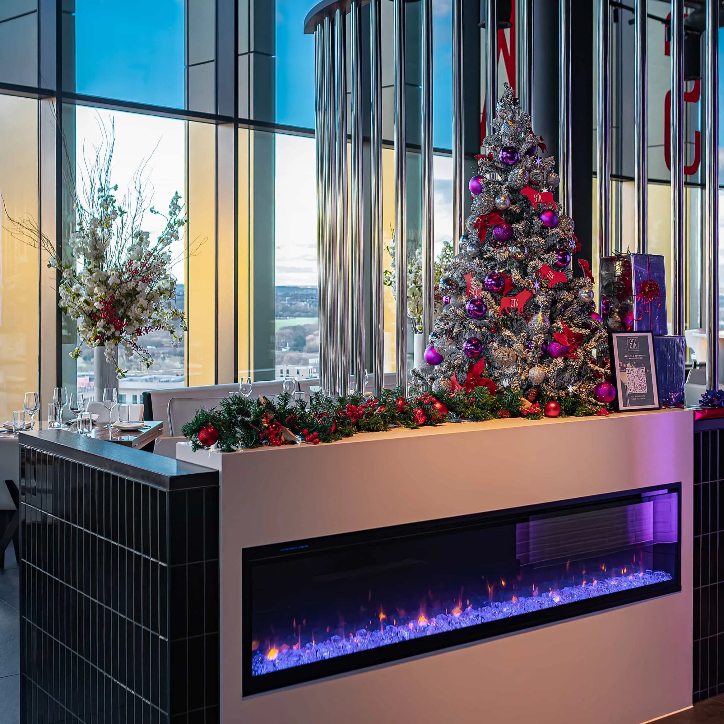 Warmly lit dining space at STK Steakhouse featuring a festive Christmas tree adorned with purple and red decorations and a modern electric fireplace, with a scenic city backdrop visible through expansive windows.
