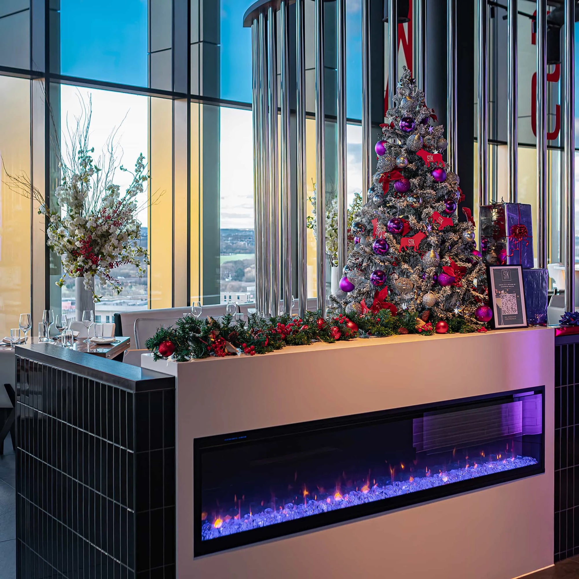 Warmly lit dining space at STK Steakhouse in London, featuring a festive Christmas tree adorned with purple and red decorations, a modern electric fireplace, and a scenic city backdrop visible through the expansive windows of this exclusive location - Amaranté London.