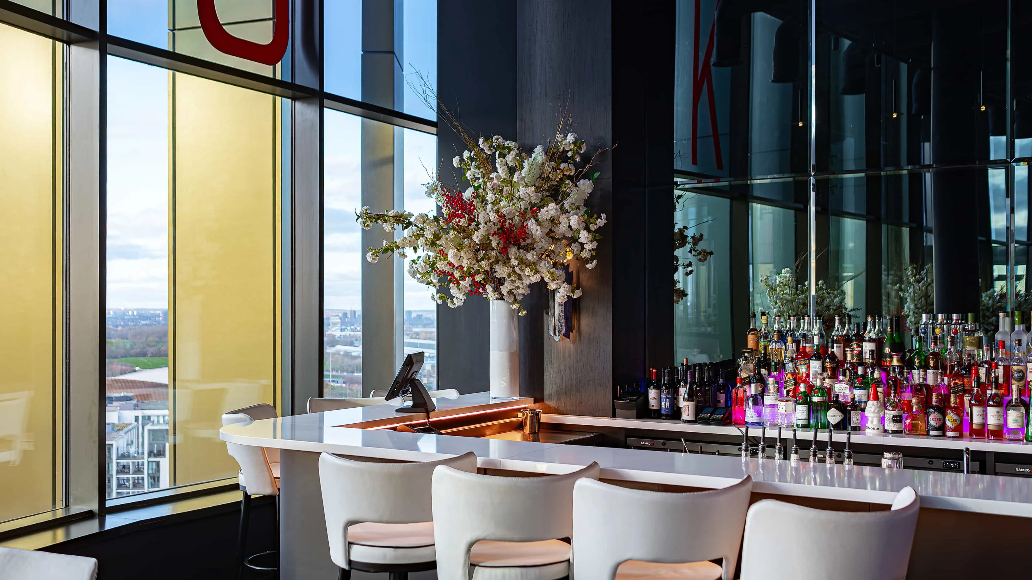 A chic bar area at STK Steakhouse with a stylish floral arrangement in a white vase, overlooking a city landscape through large windows, alongside modern furniture and a well-stocked bar.
