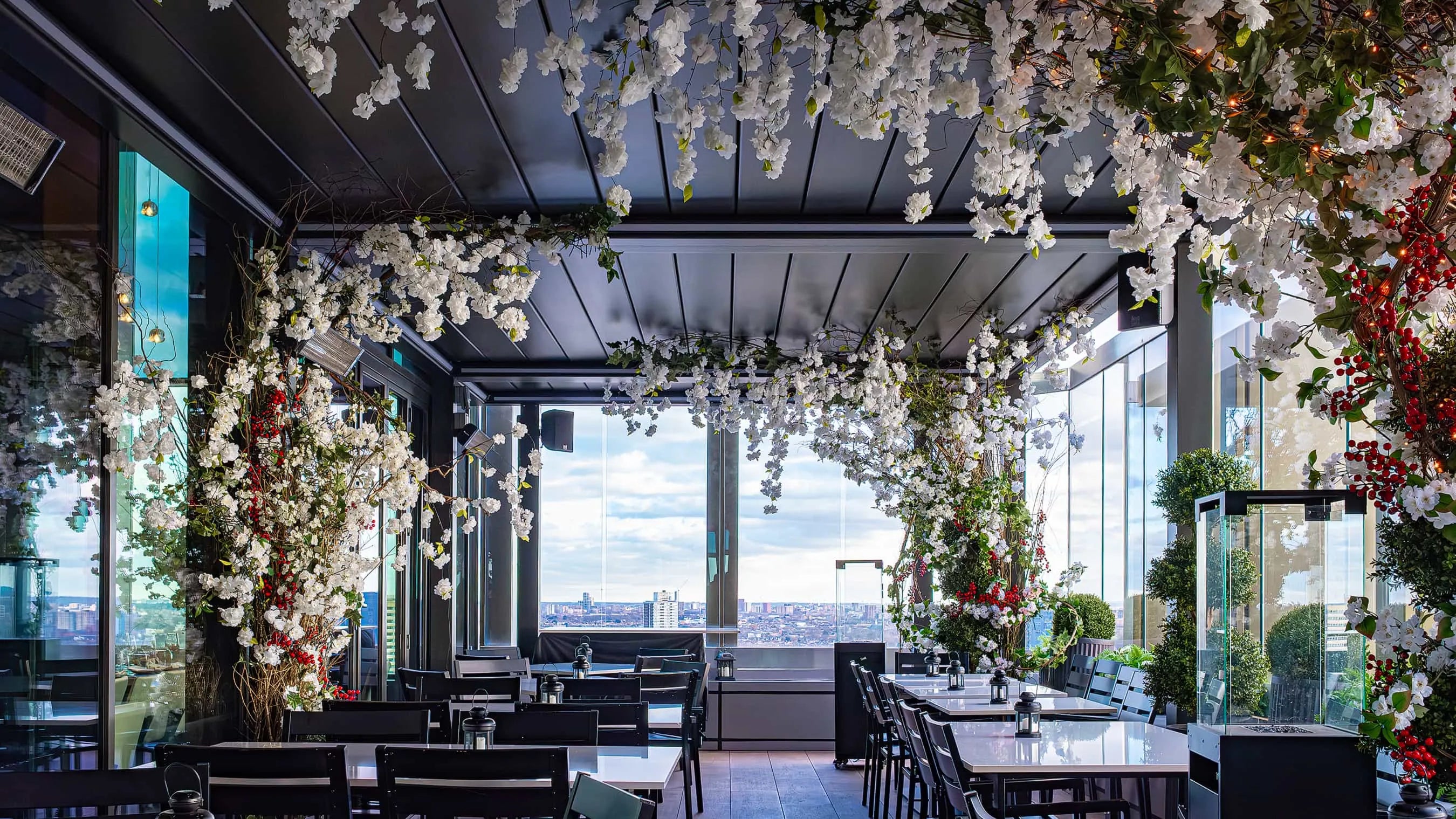 Elegant rooftop dining area at STK Steakhouse with a ceiling and pillars adorned with white blossoms and greenery, accented with red berries, offering a panoramic view of the cityscape through floor-to-ceiling windows.
