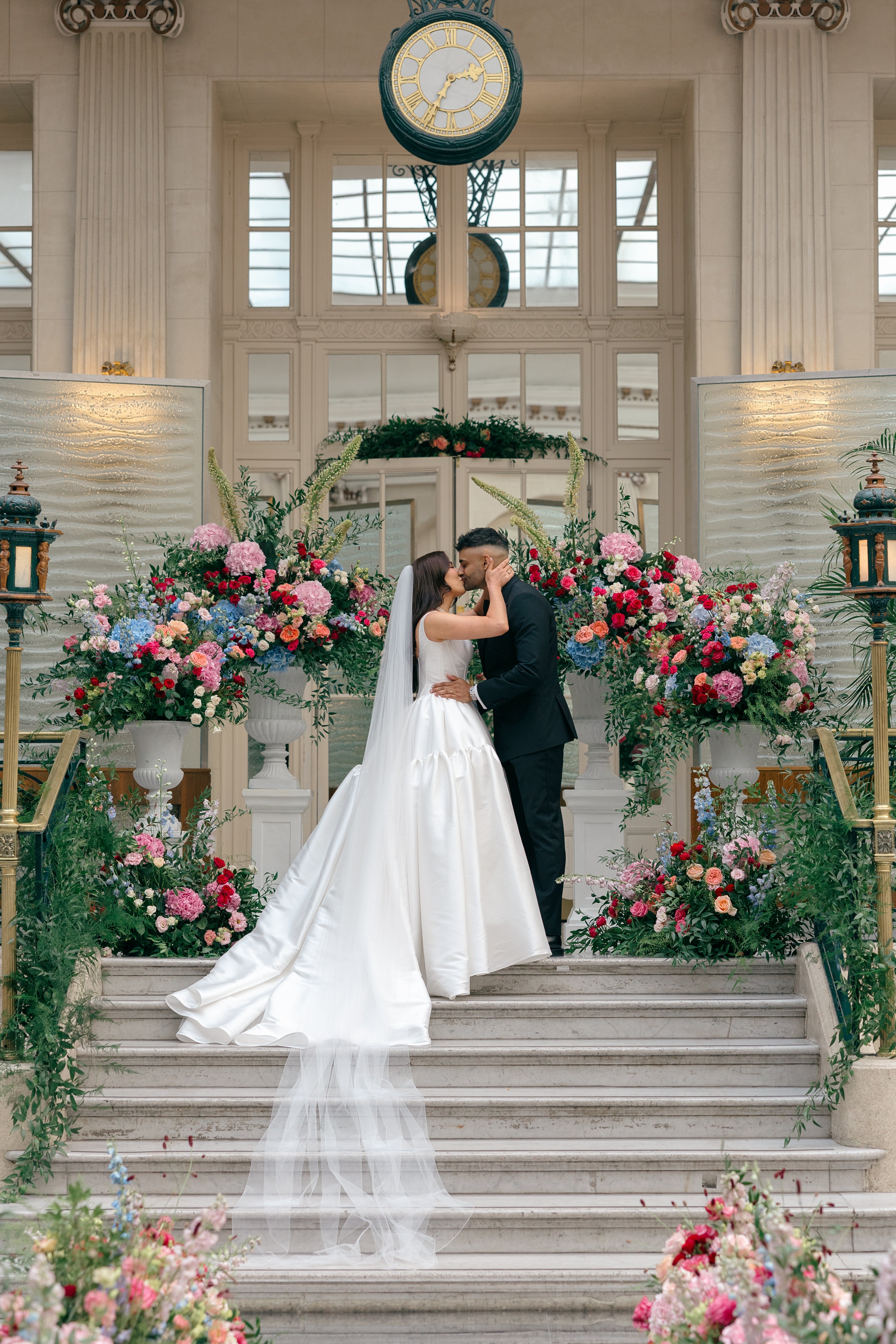 Elaborate bespoke wedding floral arrangements featuring vibrant mixed blooms cascading down a classical staircase with coordinated urn displays in a grand luxury venue, floral design by Amaranté London