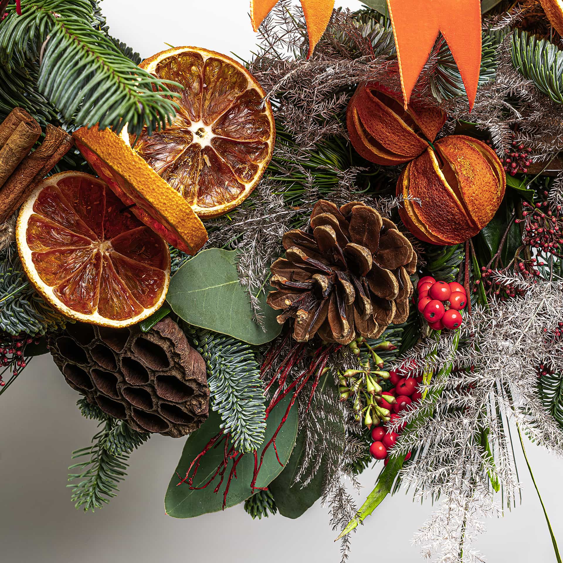 Macro detail of luxury Christmas wreath featuring dried orange slices with golden rims, brown textured pinecones, fresh dark green conifer sprigs, bright red berry clusters on crimson stems, fresh eucalyptus leaves, grey ornamental dried grasses, and coral-orange ribbon accent - Amaranté London