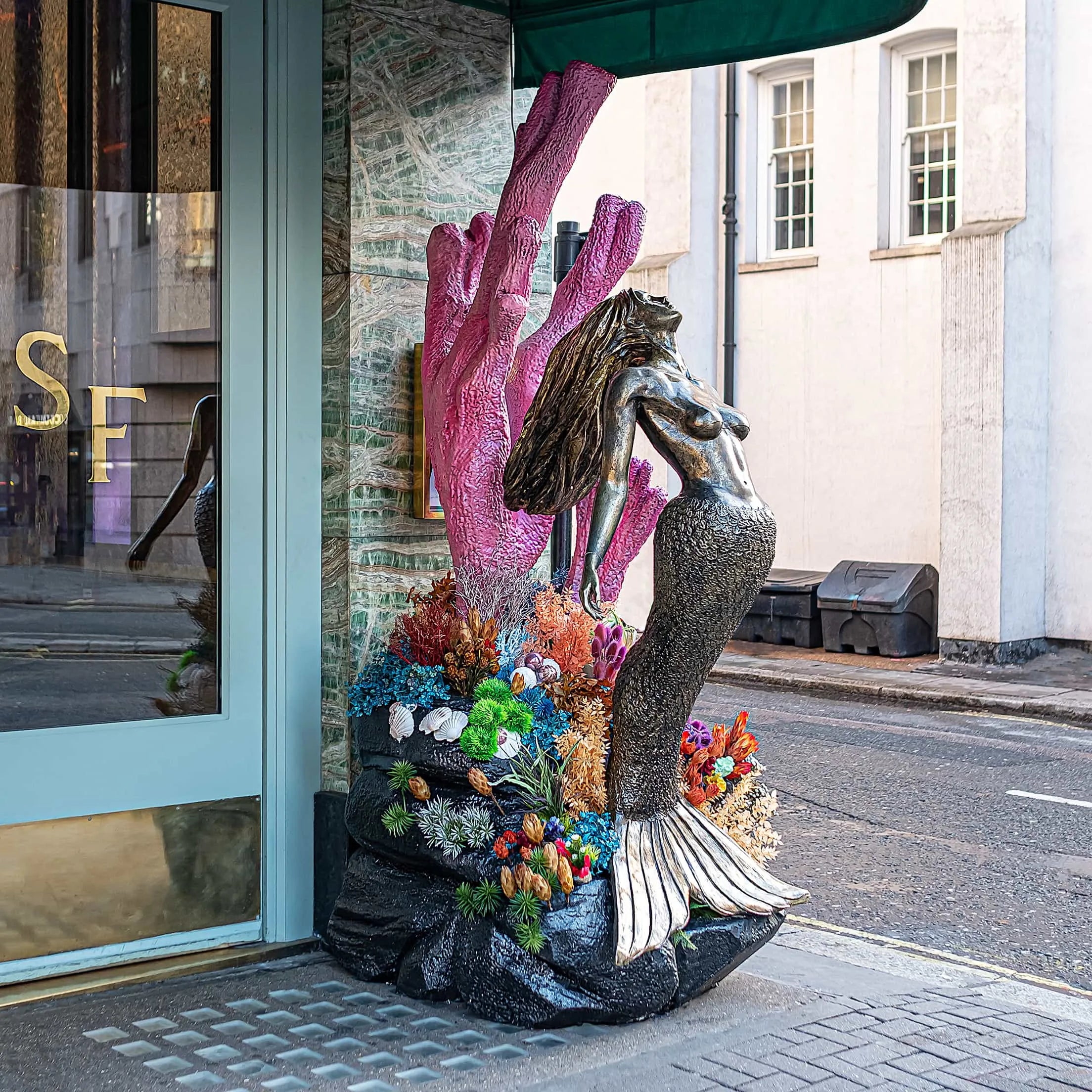 Elegant Christmas floral arrangement by Amaranté London at Sexy Fish Restaurant's entrance, featuring a mermaid statue surrounded by vibrant pink and blue coral-like stems, green ferns, and colourful flowers, creating an oceanic festive atmosphere.