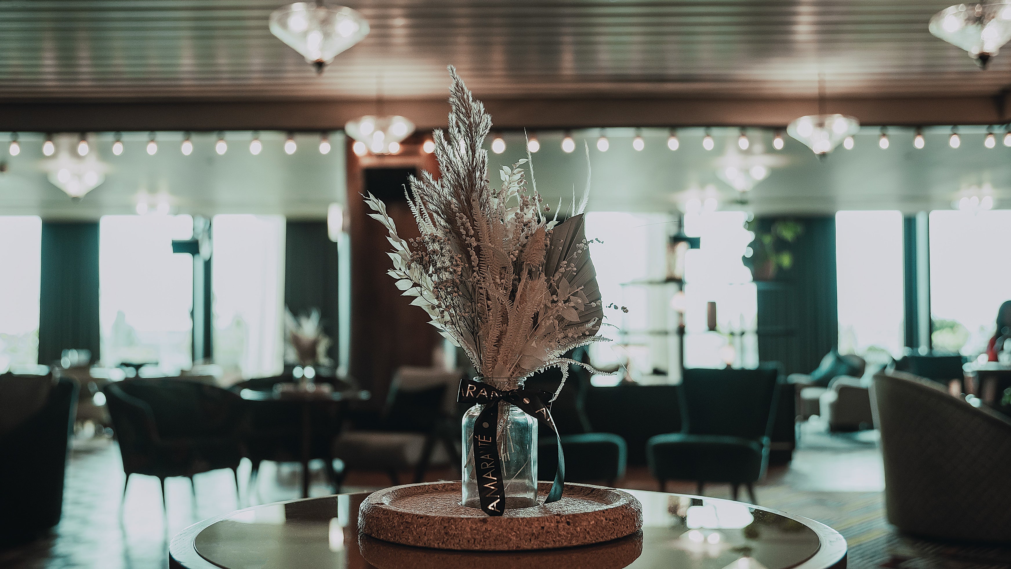 In the foreground, an elegant cork-based centrepiece showcasing Amaranté's bespoke floral arrangement takes centre stage, with an array of feather-like silvery leaves and delicate flowers emerging from a clear glass vase, set against the softly lit, luxurious backdrop of White City House's spacious lounge area.