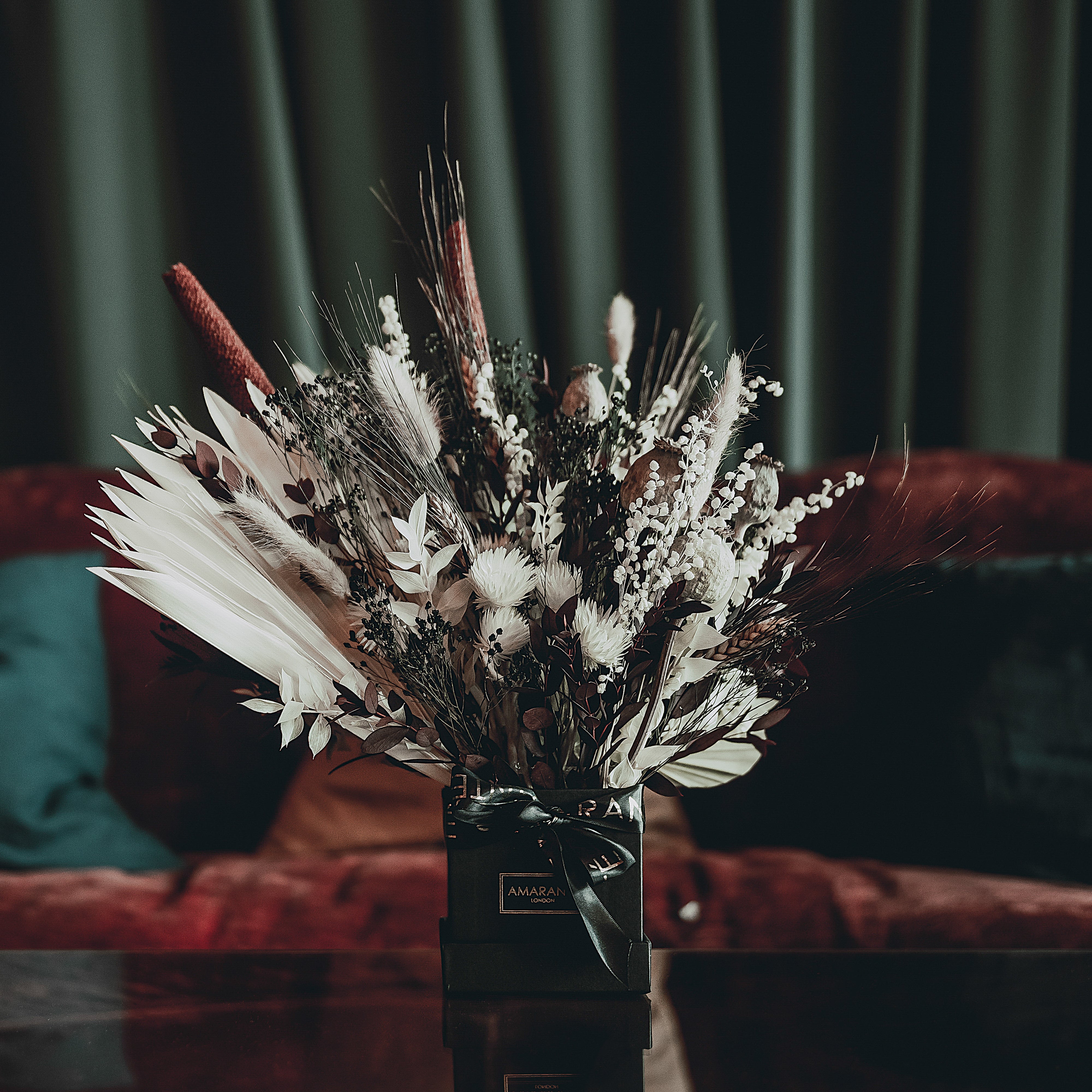 A luxurious bespoke floral arrangement by Amaranté London - Event Florist  - sits proudly in a sleek black vase with a dynamic composition of white blossoms, feathery greens, and rich red accents, providing an abundant focal point against the deep red velvet backdrop of an elegant event setting.