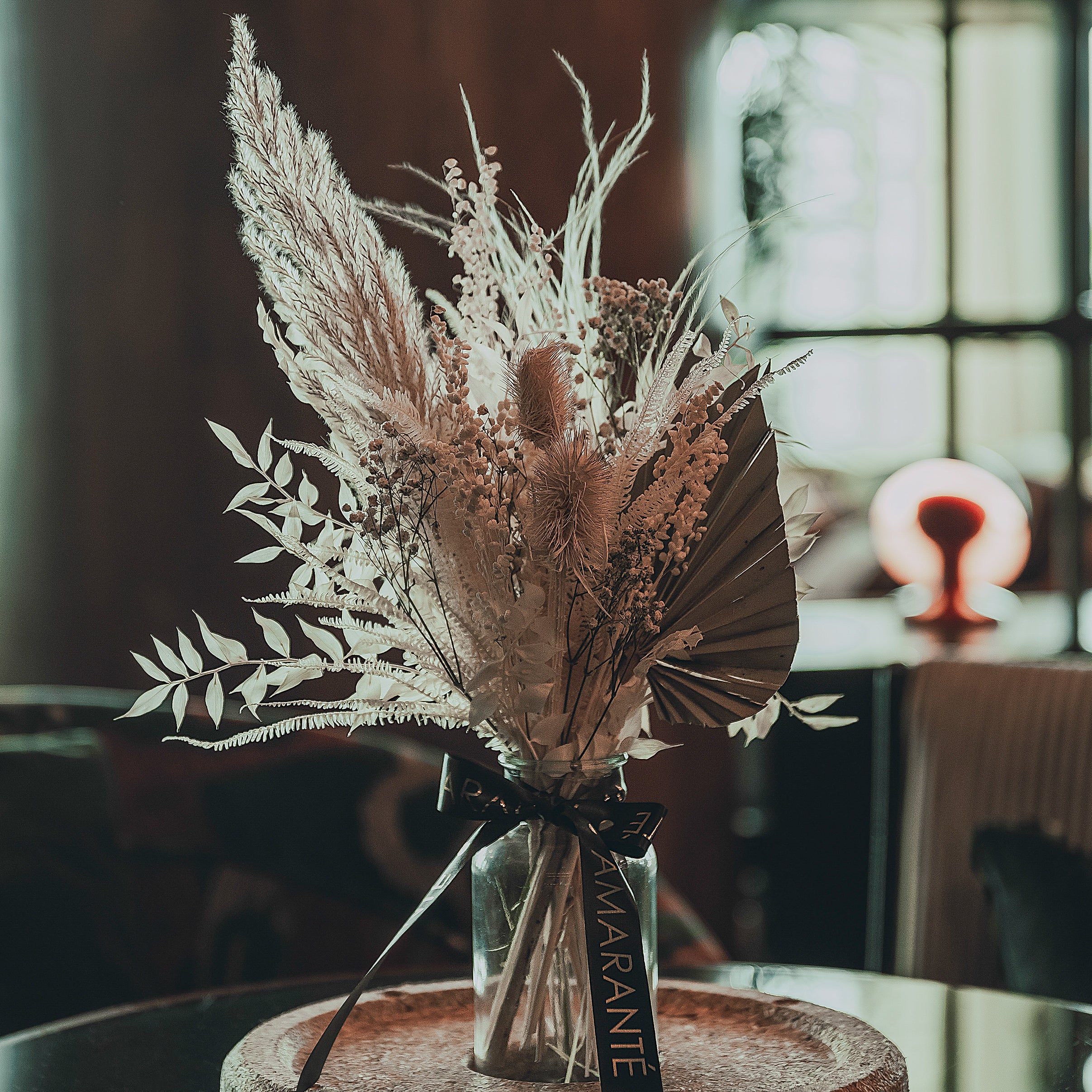 In the foreground, an elegant cork-based centrepiece showcasing Amaranté's bespoke floral arrangement takes centre stage, with an array of feather-like silvery leaves and delicate flowers emerging from a clear glass vase, set against the softly lit, luxurious backdrop of White City House's spacious lounge area.