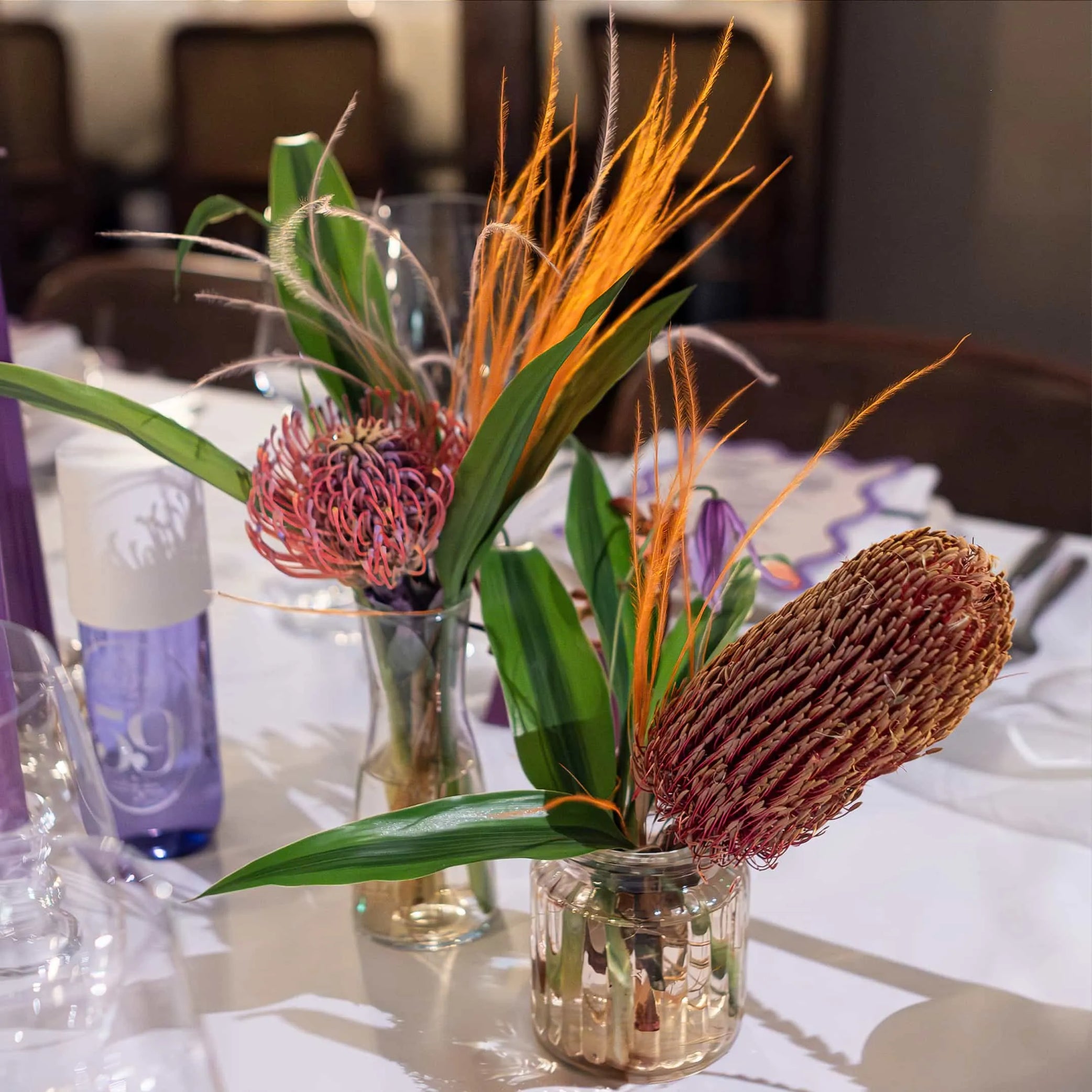 Detailed view of Amaranté London's table floral arrangements with vibrant orange feathers and exotic proteas in glass vases for Sol de Janeiro event setting.