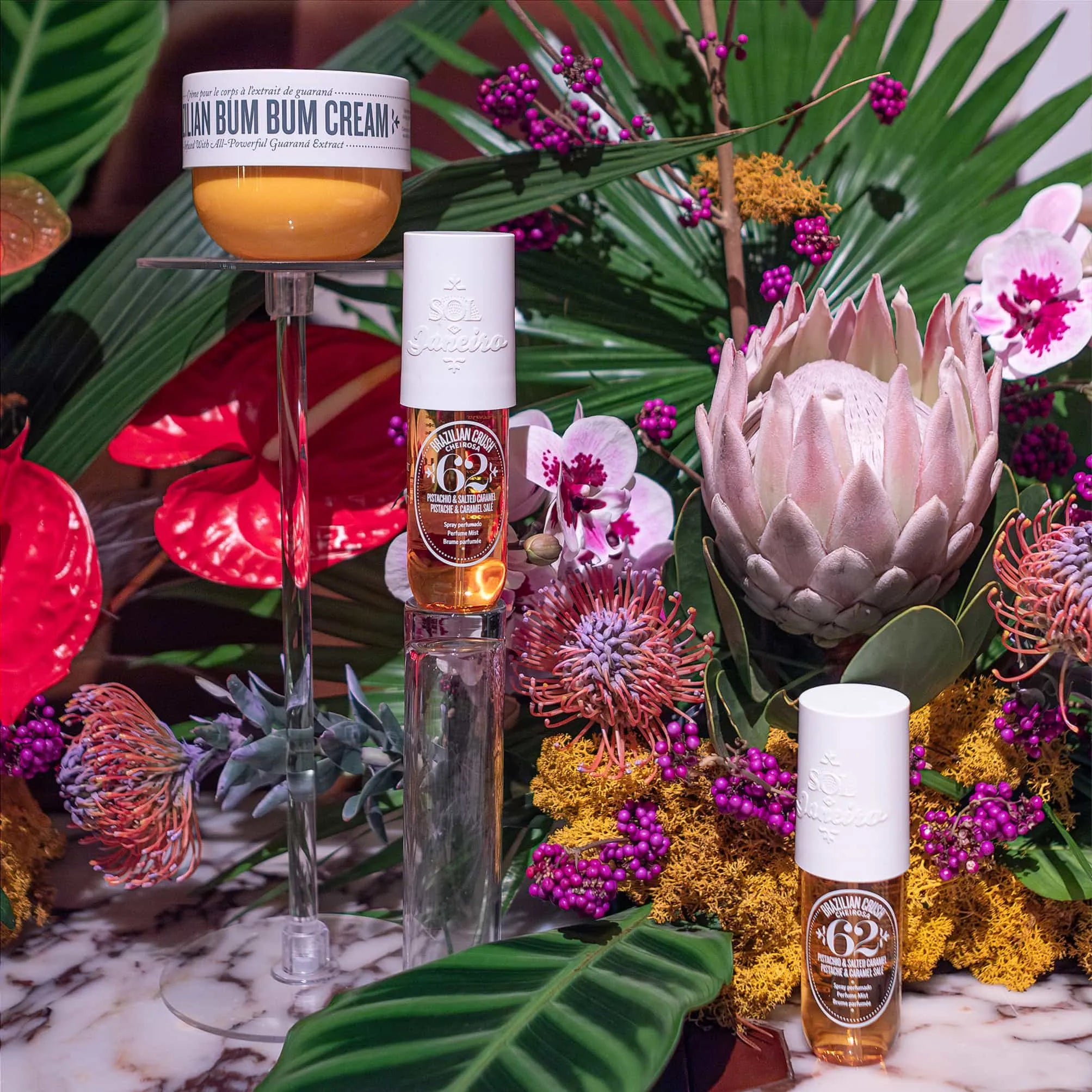 Lavish display of Sol de Janeiro beauty products, including Brazilian Bum Bum Cream and Glow Oils, set among a vibrant arrangement of pink orchids, proteas, and tropical flora by Amaranté London.