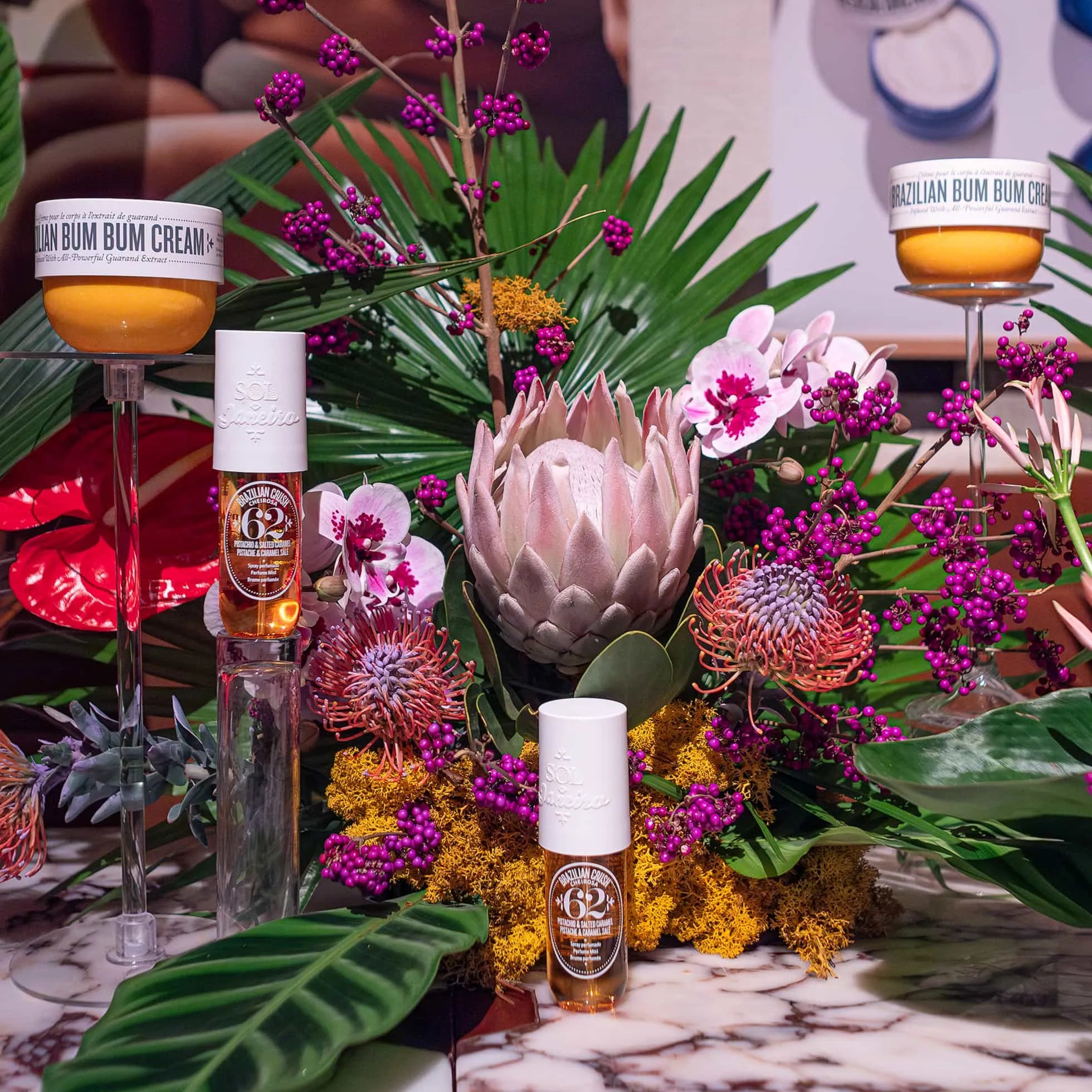 Close-up of Sol de Janeiro's Brazilian Bum Bum Cream and body spray elegantly displayed among a rich array of Amaranté London's floral arrangements with orchids, proteas, and tropical leaves.