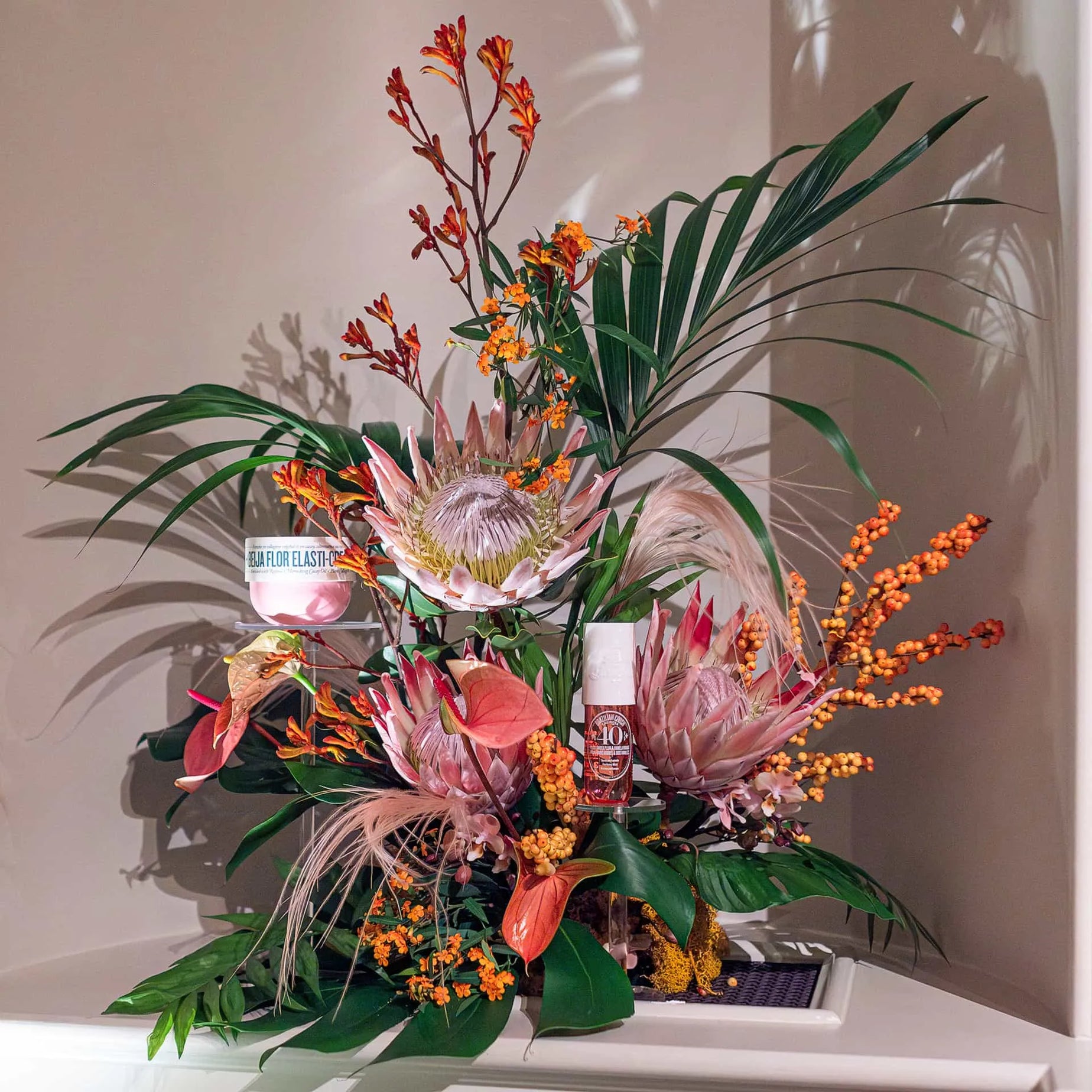 Amaranté London's intricate floral arrangement featuring a mix of orange blossoms, proteas, and tropical leaves, showcasing Sol de Janeiro's Beija Flor Elasti-Cream against a soft, shadowed backdrop.