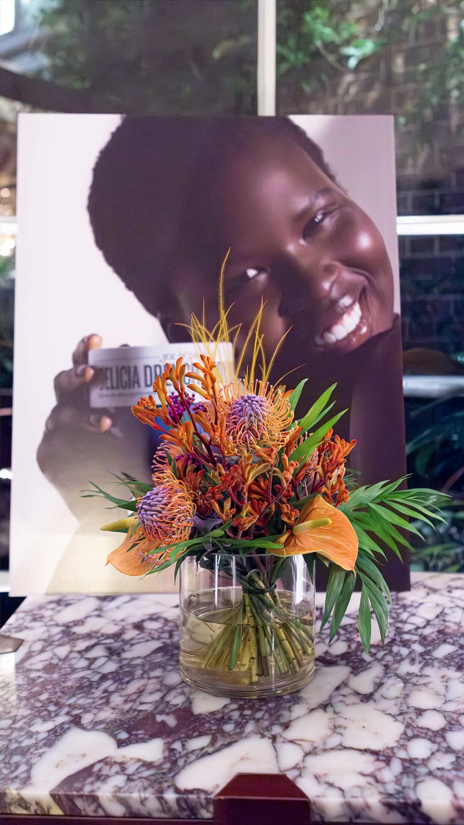 Colorful floral arrangement by Amaranté London in a clear vase on a marble table, complementing the joyful theme of Sol de Janeiro's advertisement in the background.