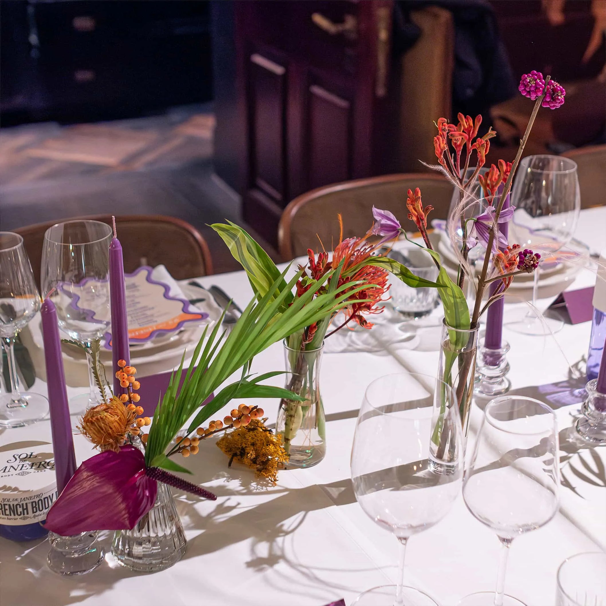 Sophisticated table setting with Amaranté London's floral decorations, featuring slender vases with tropical blooms and candles, prepared for a Sol de Janeiro launch event.