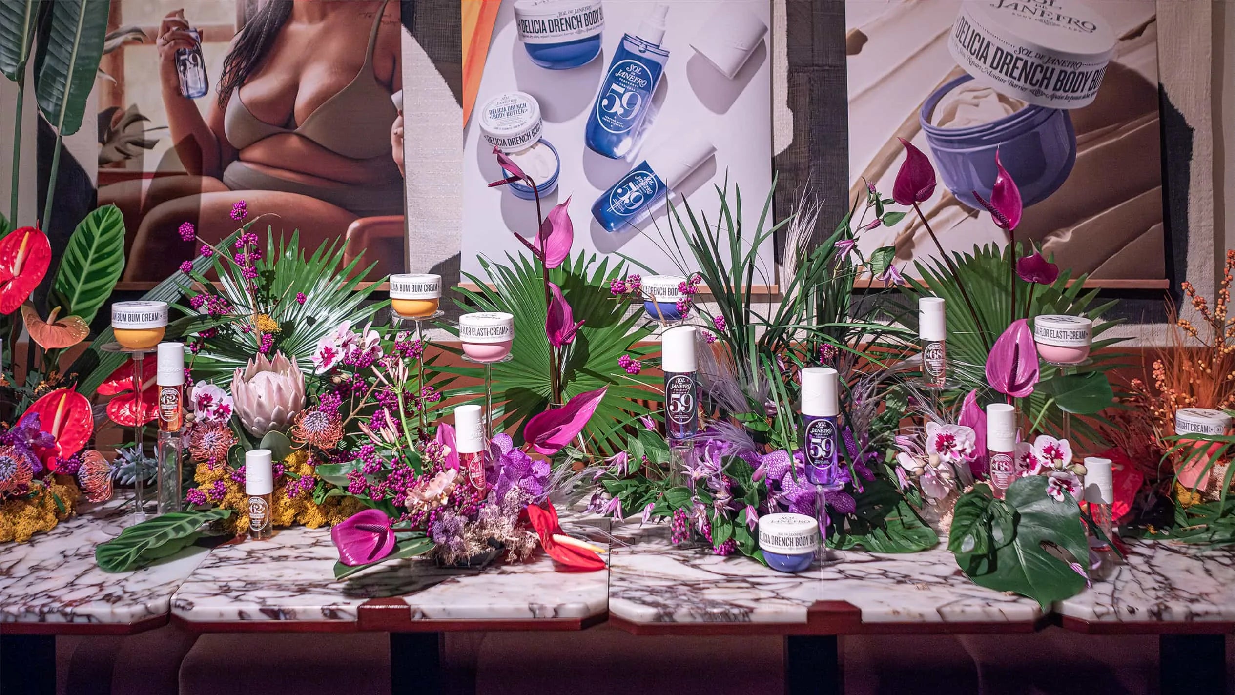 A lavish array of Sol de Janeiro's beauty products elegantly presented on a grand table amid Amaranté London's striking floral arrangements, with a backdrop of chic interior decor at Maine Mayfair restaurant.